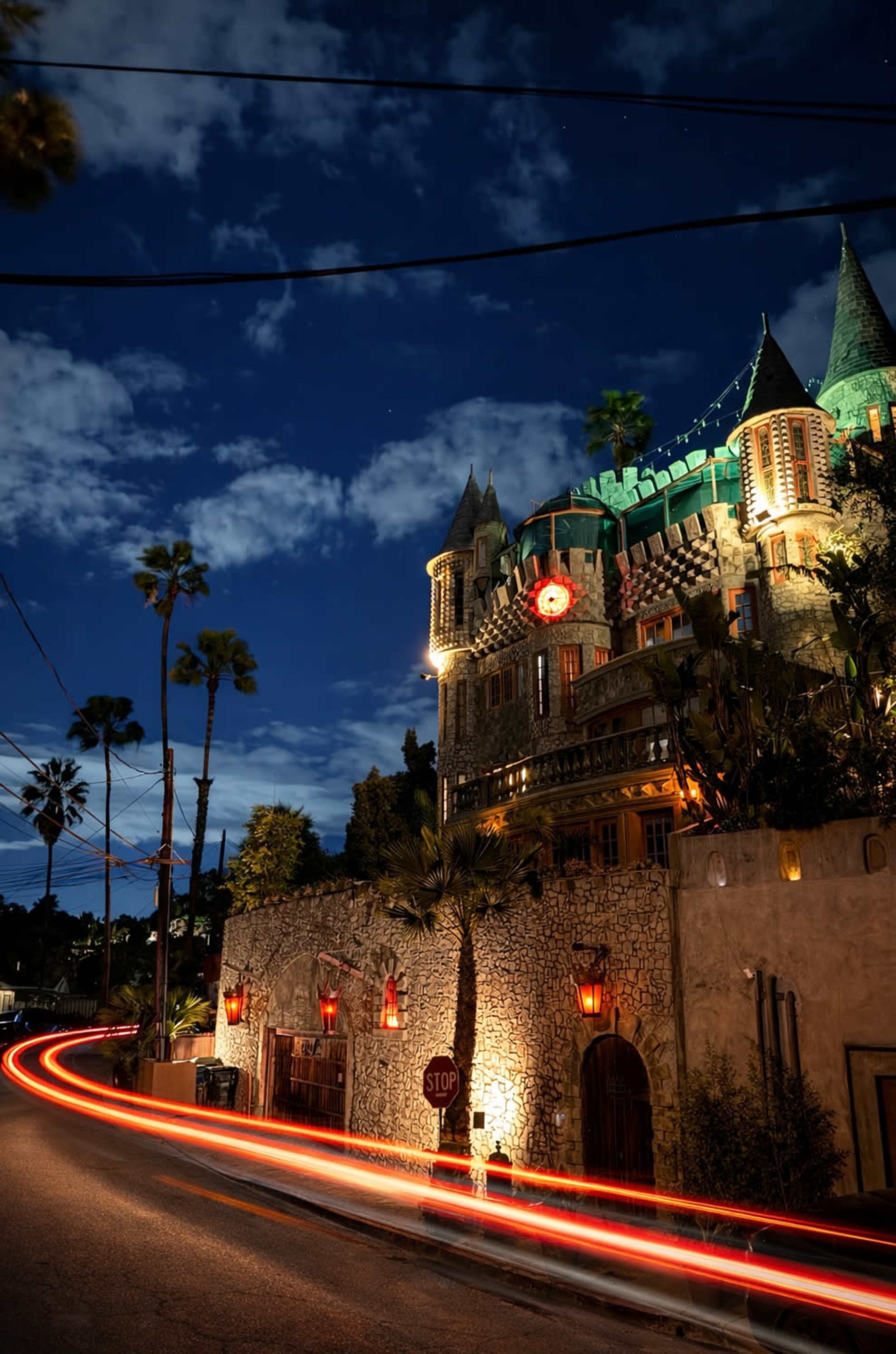 A stone castle with turrets and green roofs is illuminated at night, surrounded by palm trees and a winding road marked with a stop sign.