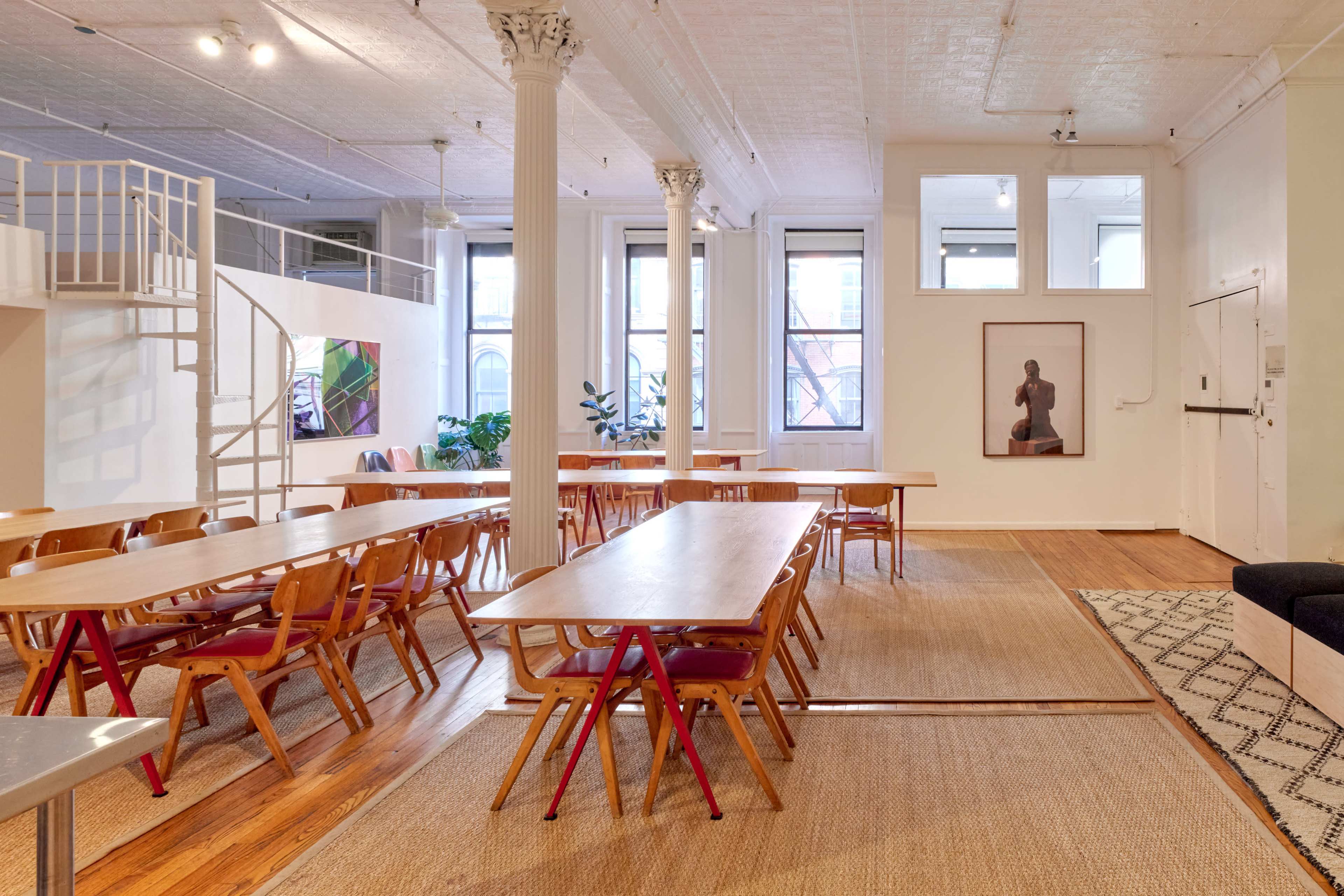 The image shows a spacious, modern interior with several long tables and chairs arranged in a communal area, surrounded by large windows and minimalist decor.