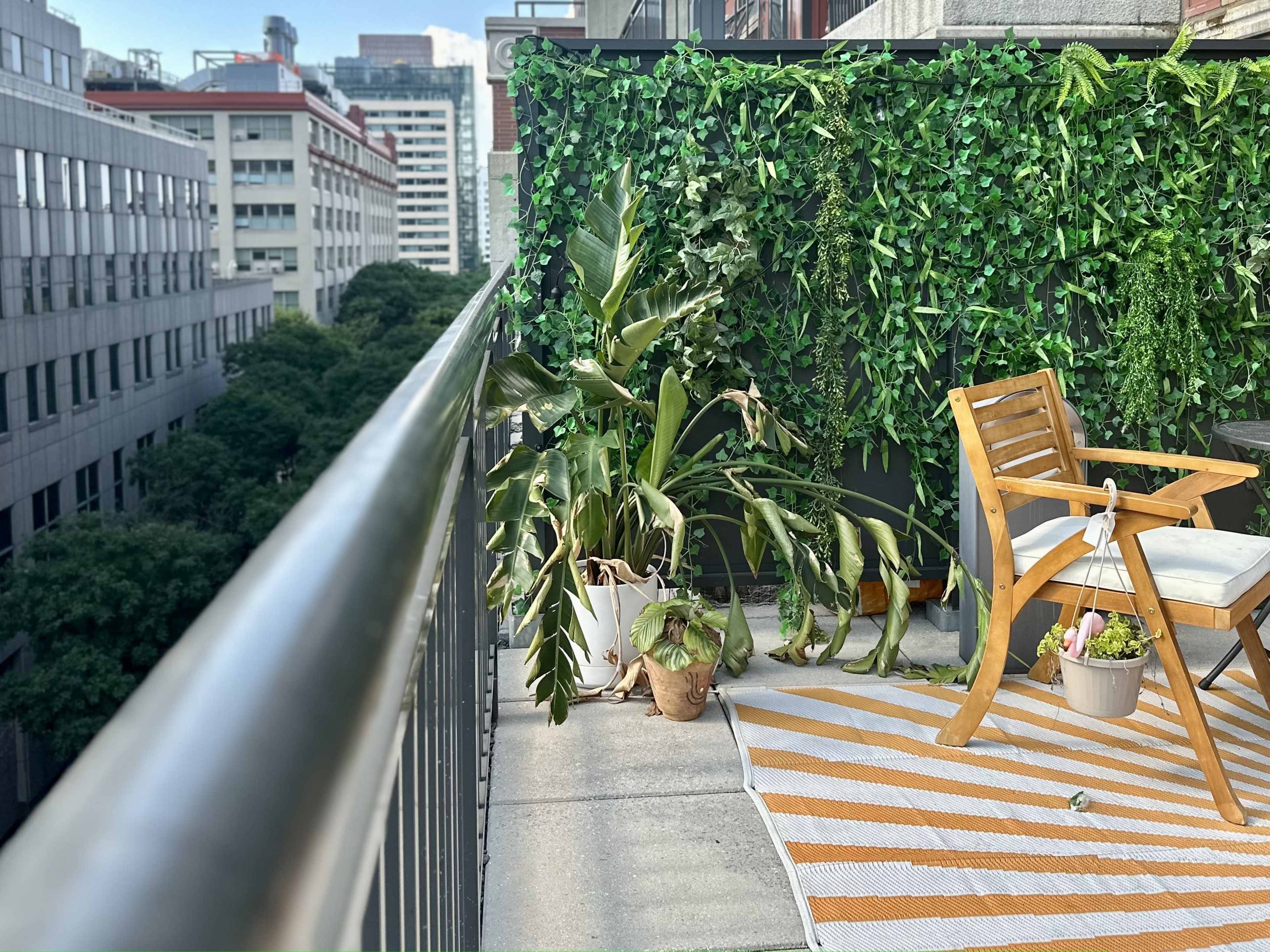 A balcony with a wooden chair, a small table with plants, and a striped rug, surrounded by greenery and tall buildings in the background.