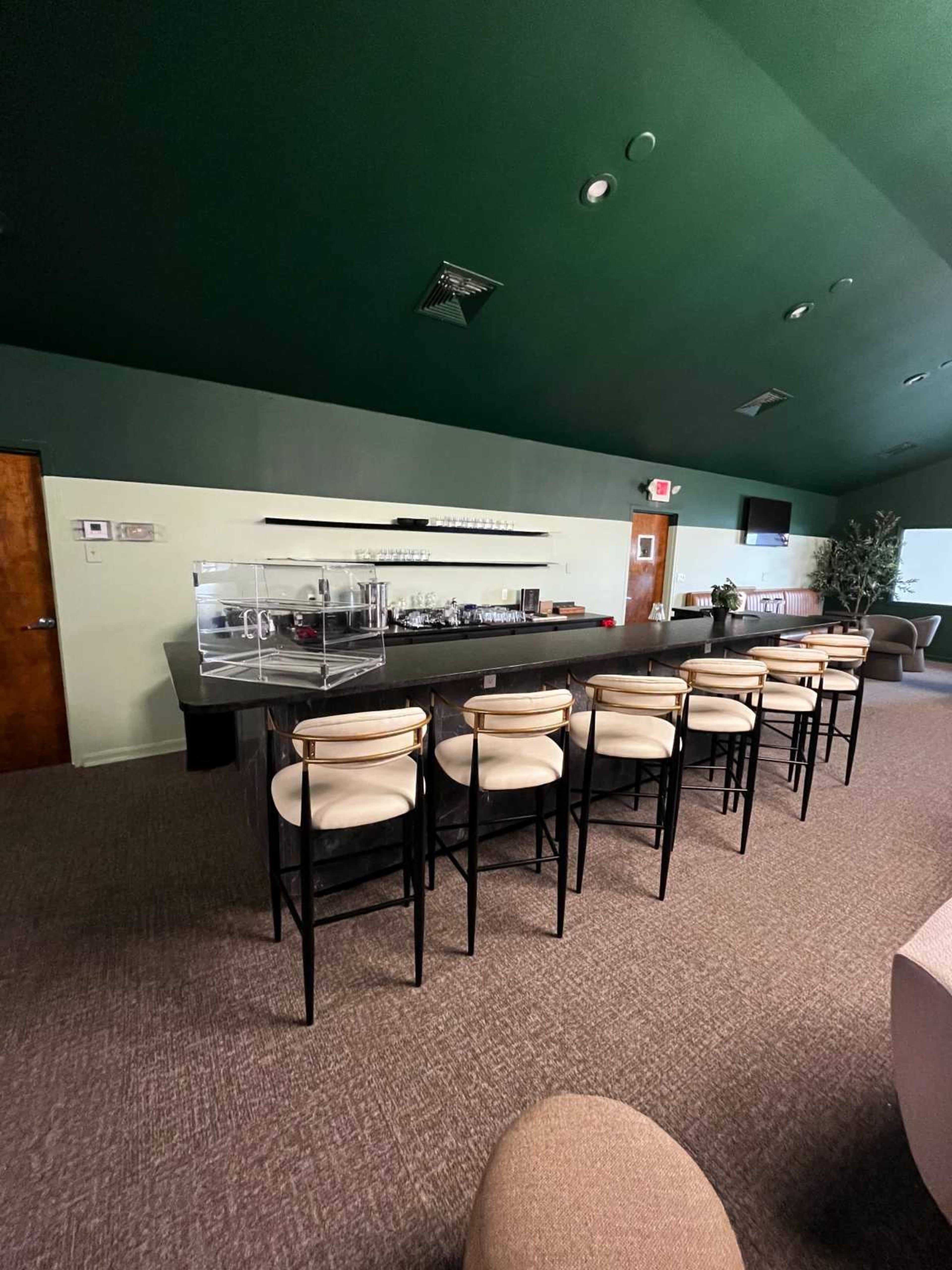The image shows a modern lounge area featuring a long bar with high stools, a green ceiling, and a minimalist decor approach.