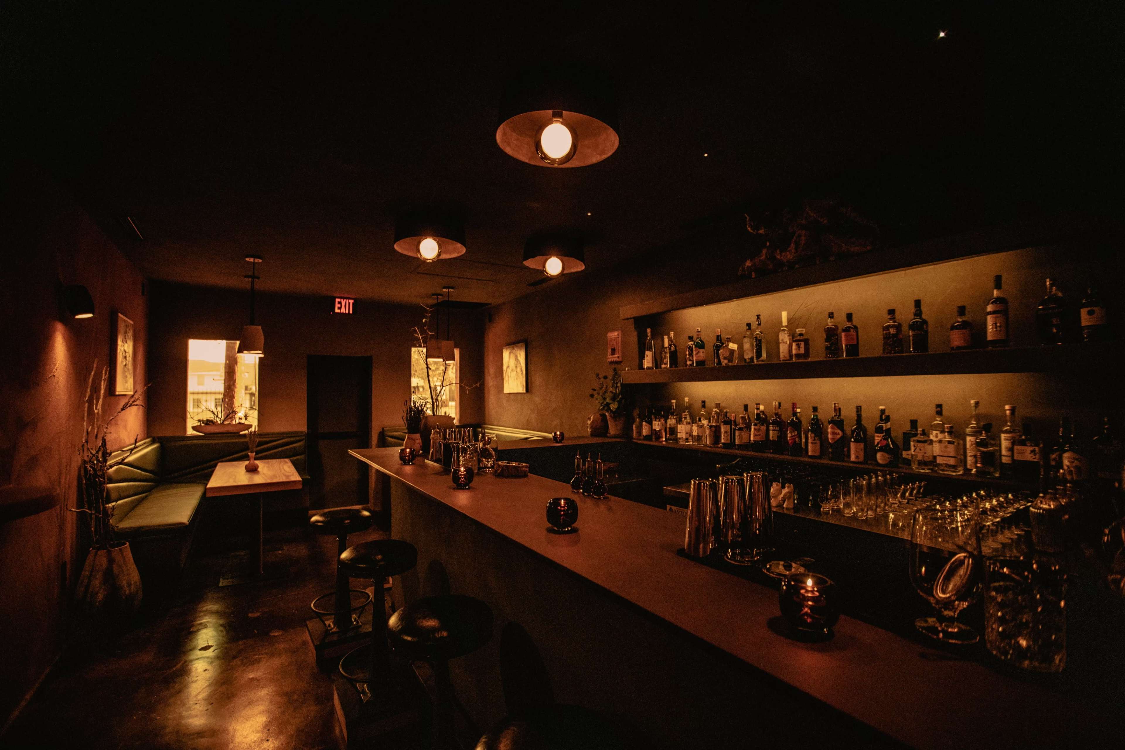 A dimly lit bar with a long counter, shelves lined with bottles, and a cozy seating area.
