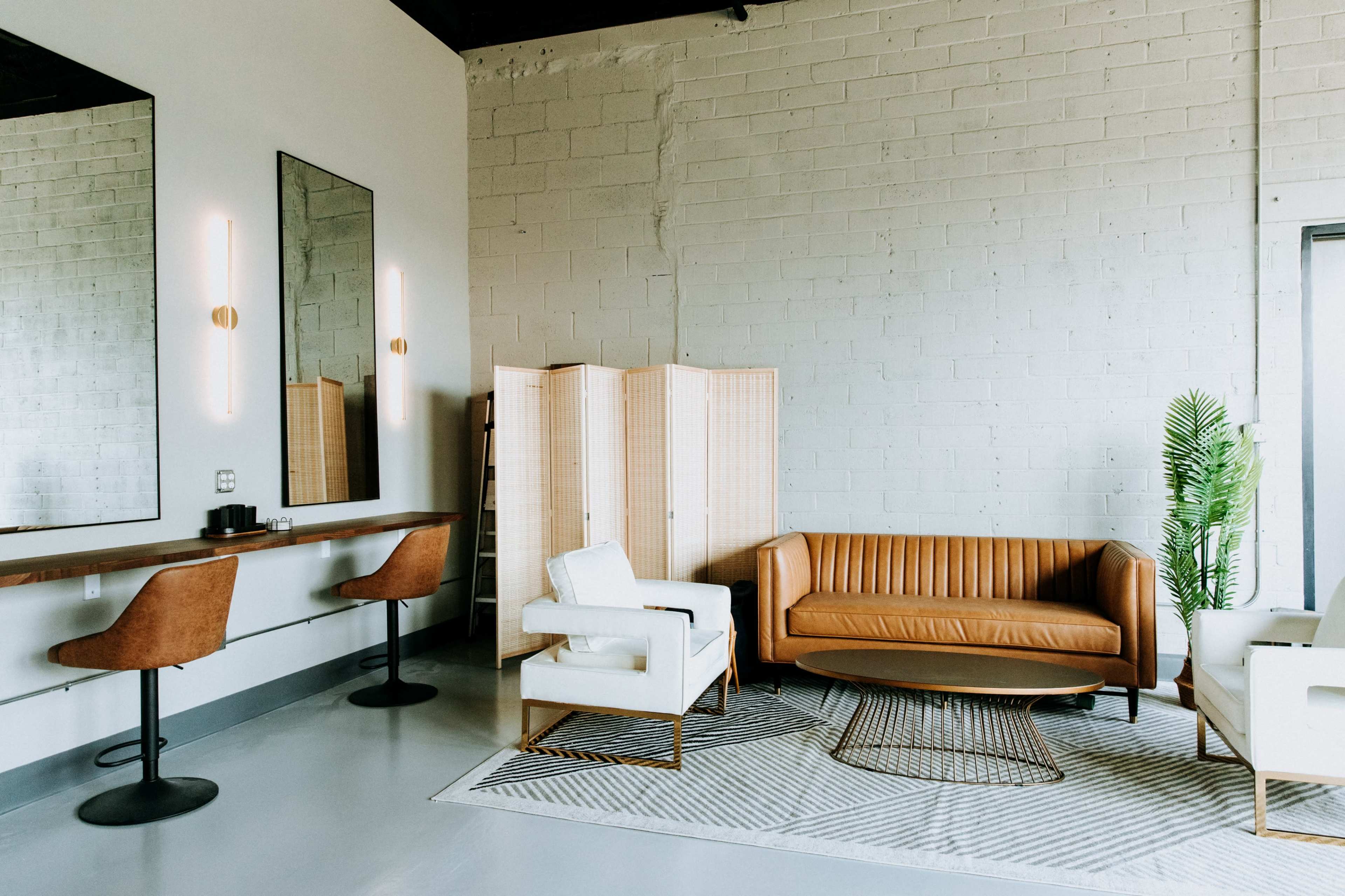 A modern living space featuring a brown leather sofa, a white armchair, a wooden table with two brown bar stools, a decorative screen, and a potted plant.