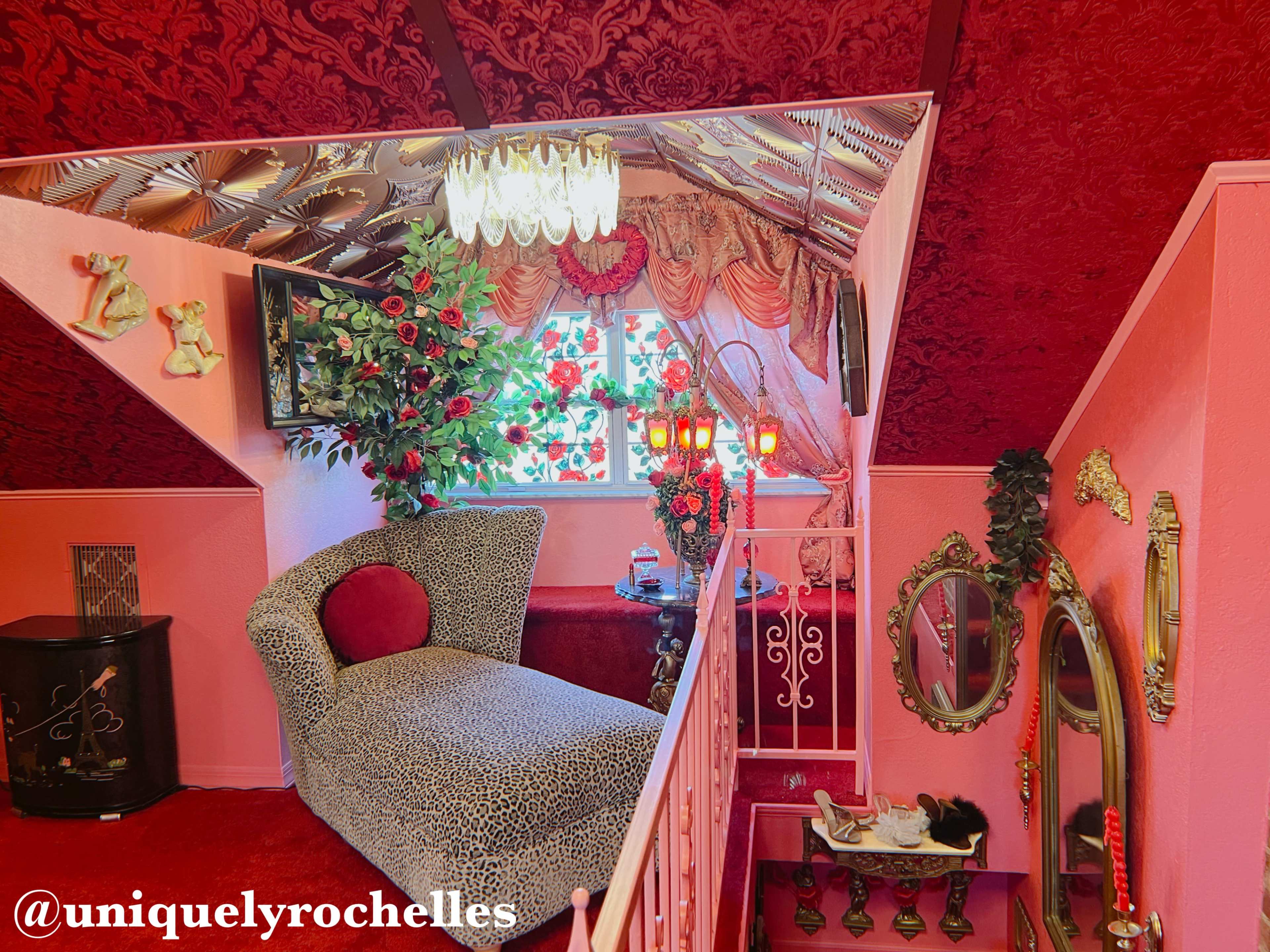 The image shows a vividly decorated corner of a room featuring a leopard-print chaise lounge, floral arrangements, ornate lighting, and rich red accents.
