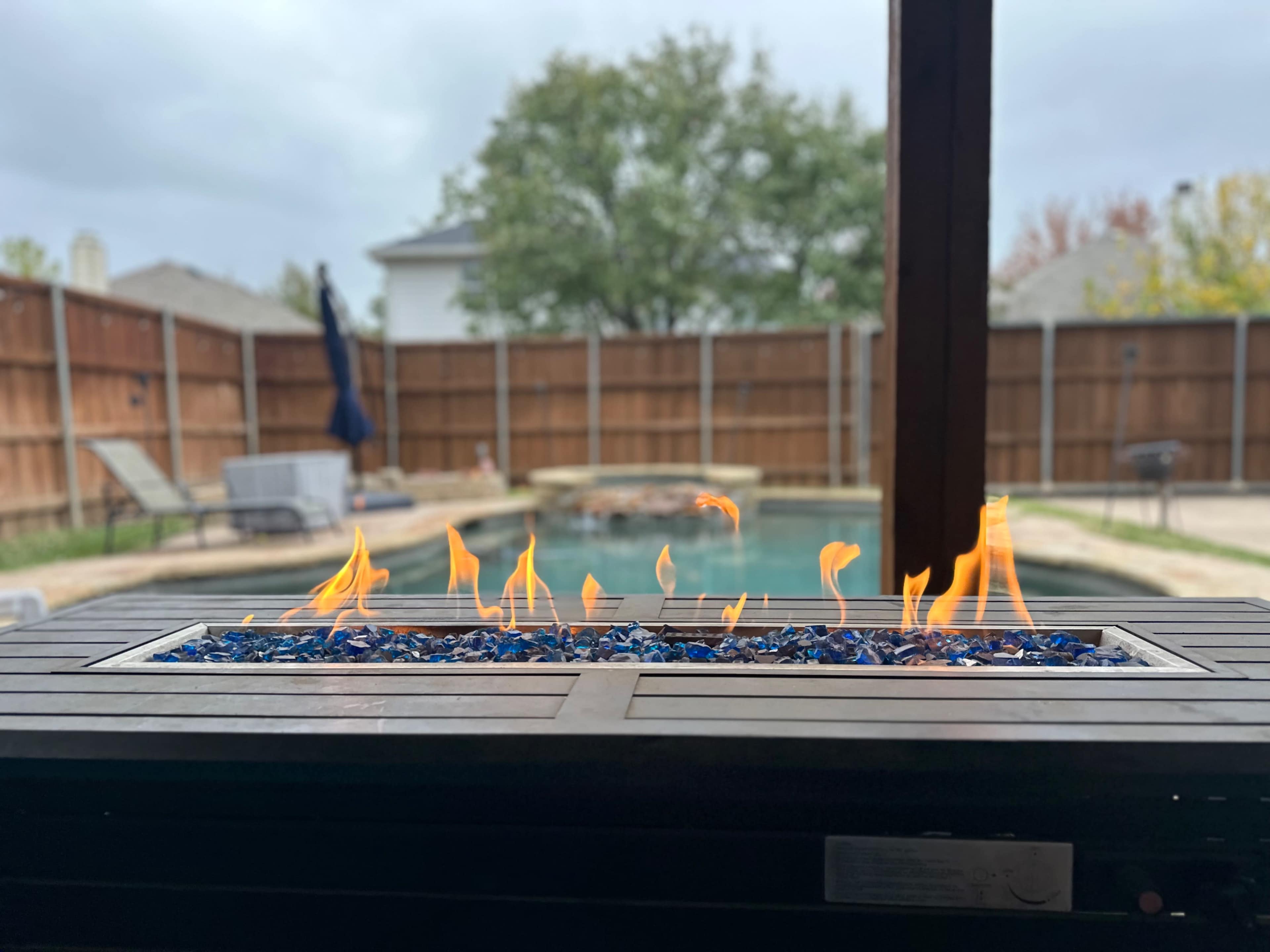 A fire pit with blue glass stones burns brightly, overlooking a swimming pool and a fenced backyard.