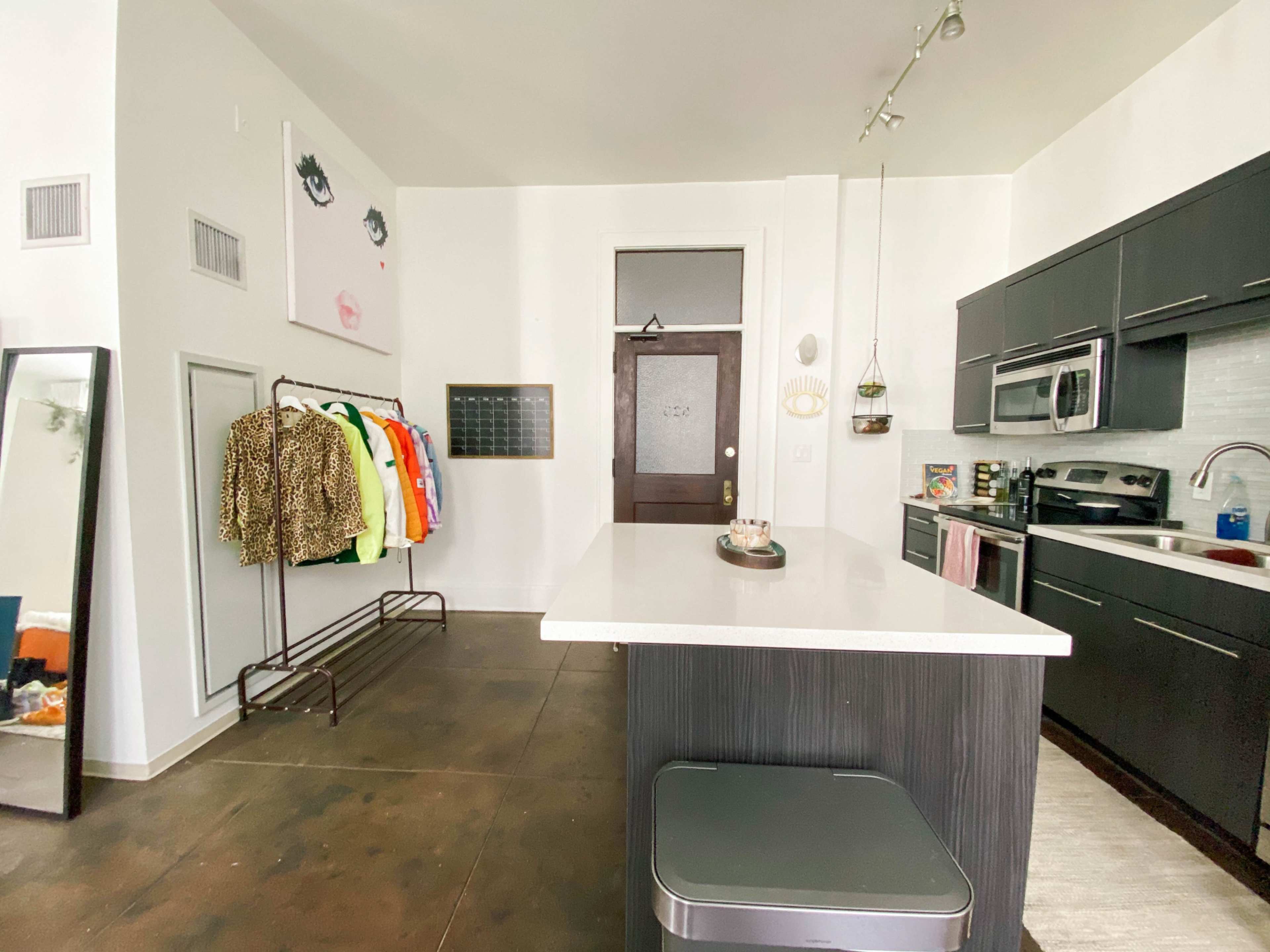 A modern kitchen with a central island, a clothing rack with various jackets, and a mirror against the wall.
