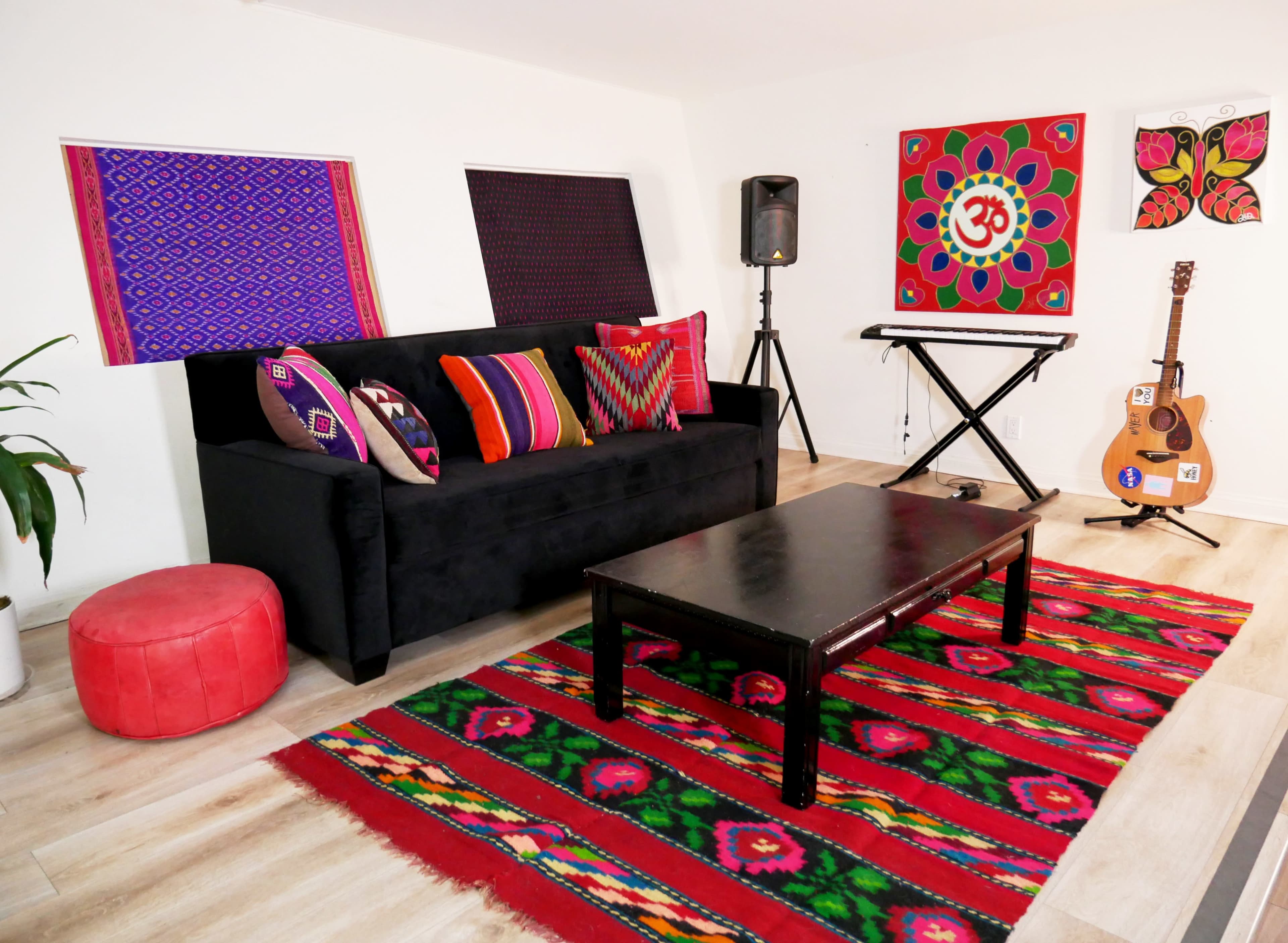 A colorful living room with a black sofa adorned with patterned cushions, a wooden coffee table, a red patterned rug, a guitar, a keyboard, and decorative wall art.