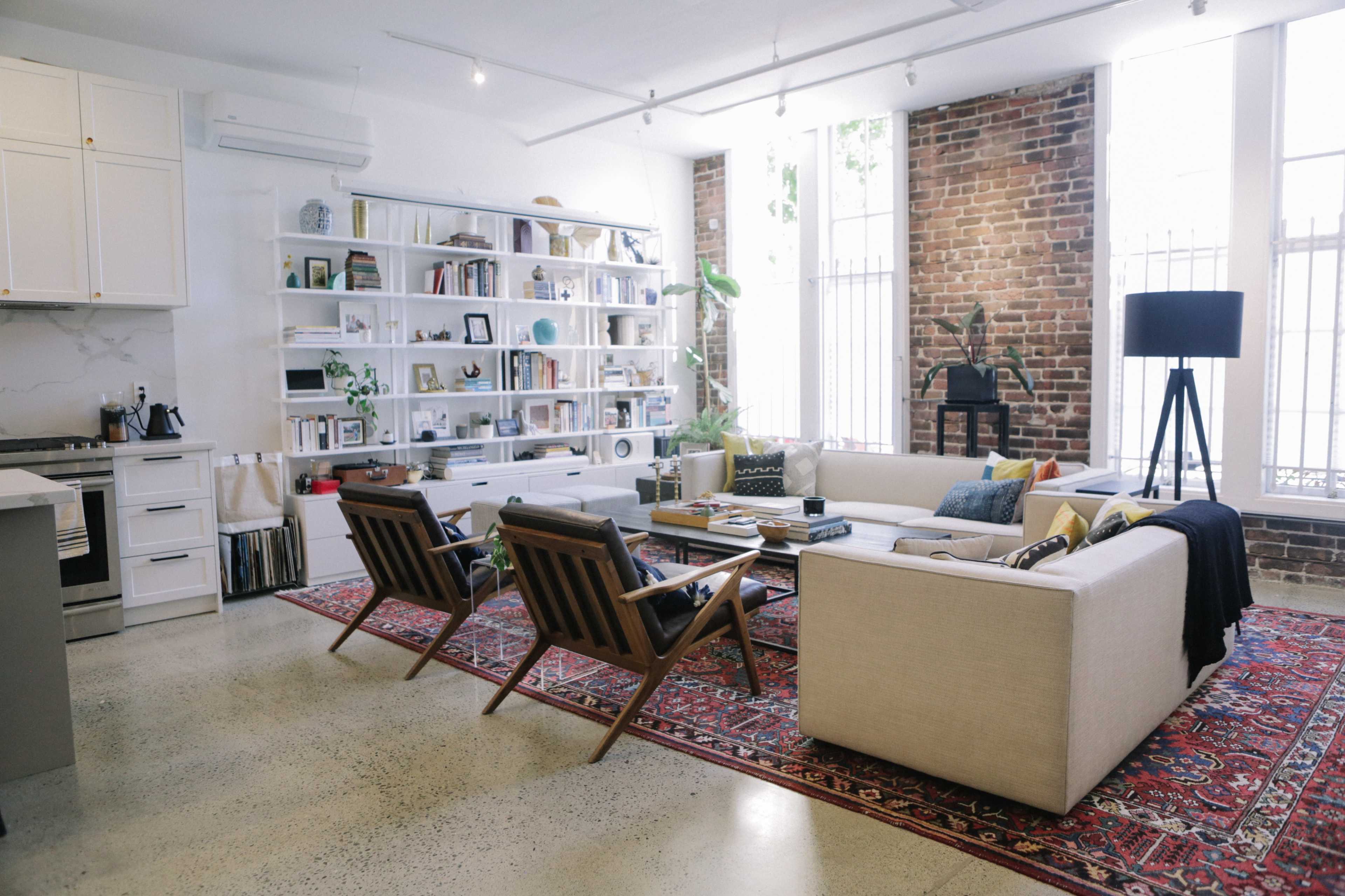A modern living room with a beige sofa, wooden chairs, a coffee table, and a bookshelf filled with books, against a backdrop of exposed brick walls and large windows.
