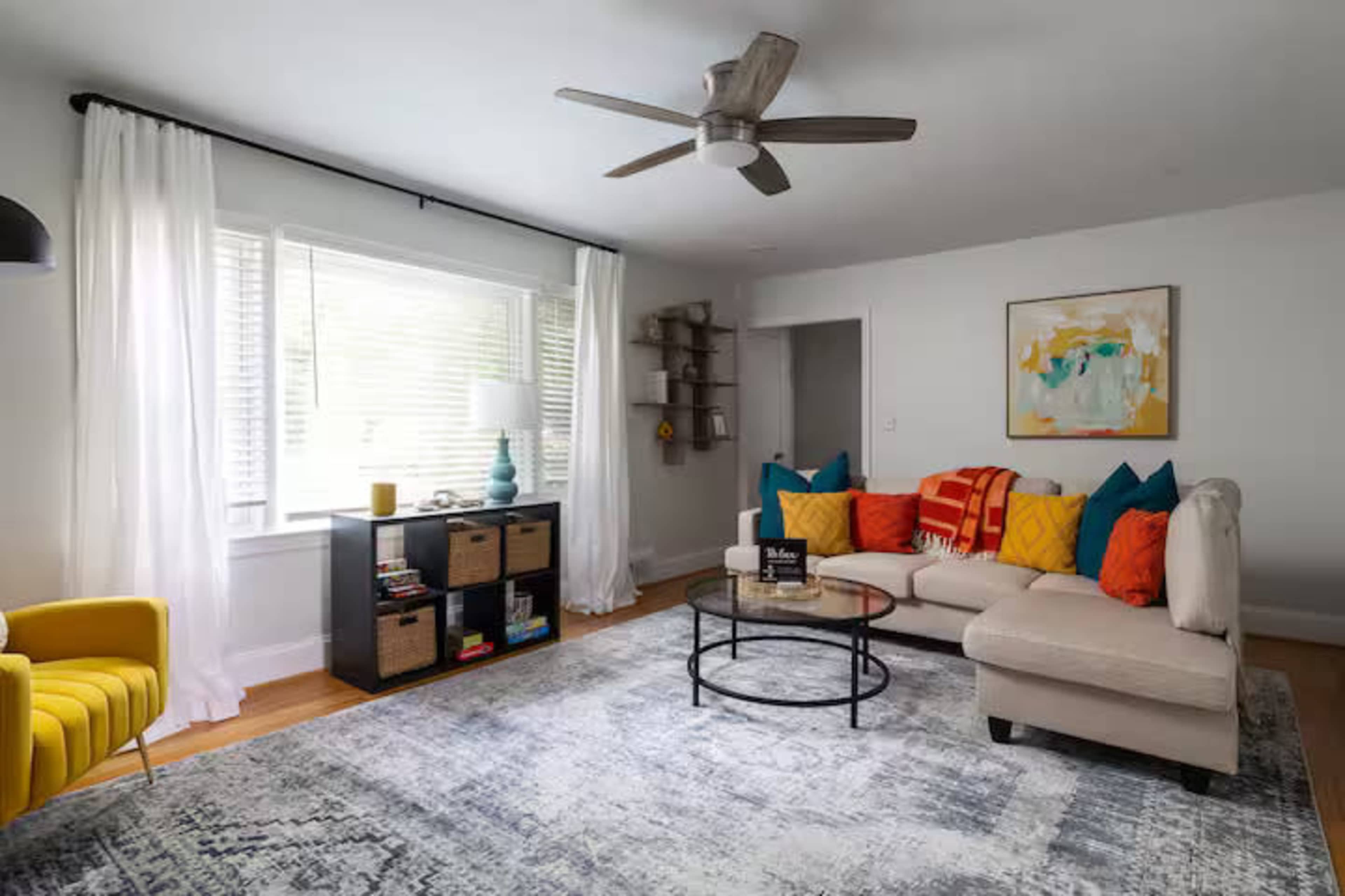 A living room features a light-colored sectional sofa with colorful pillows, a round coffee table, a yellow accent chair, and a large window with white curtains.