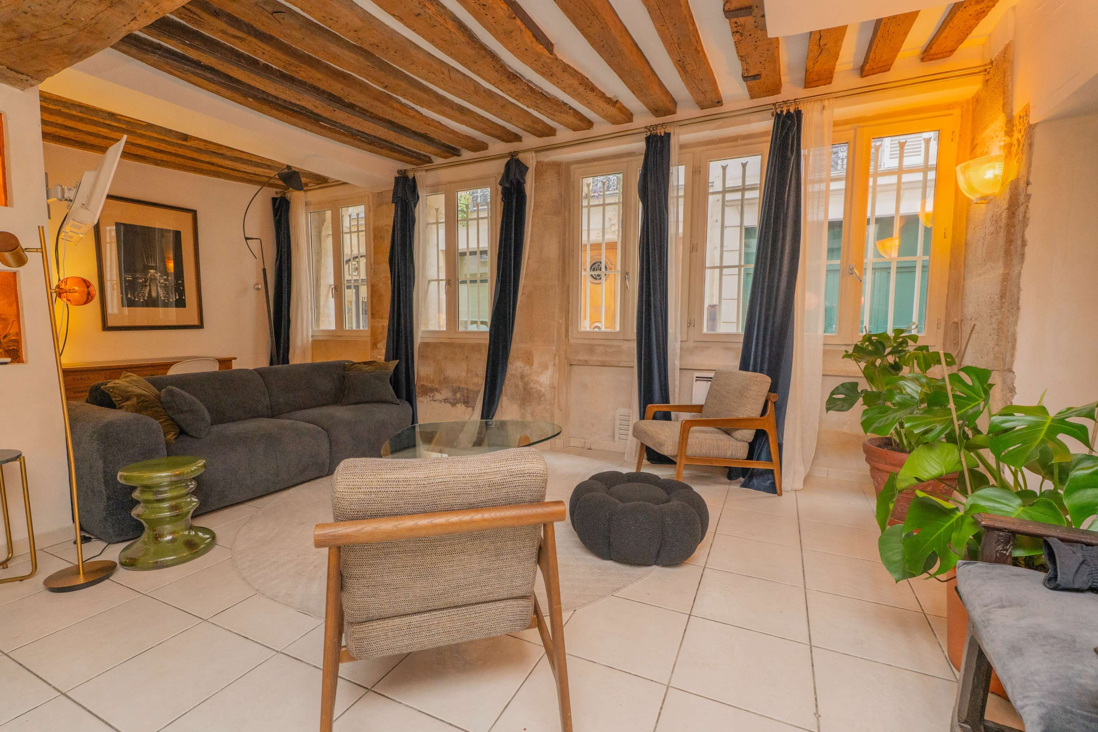 The room features exposed wooden beams, a gray sofa, a chair, and large windows with blue curtains, complemented by potted plants and ambient lighting.