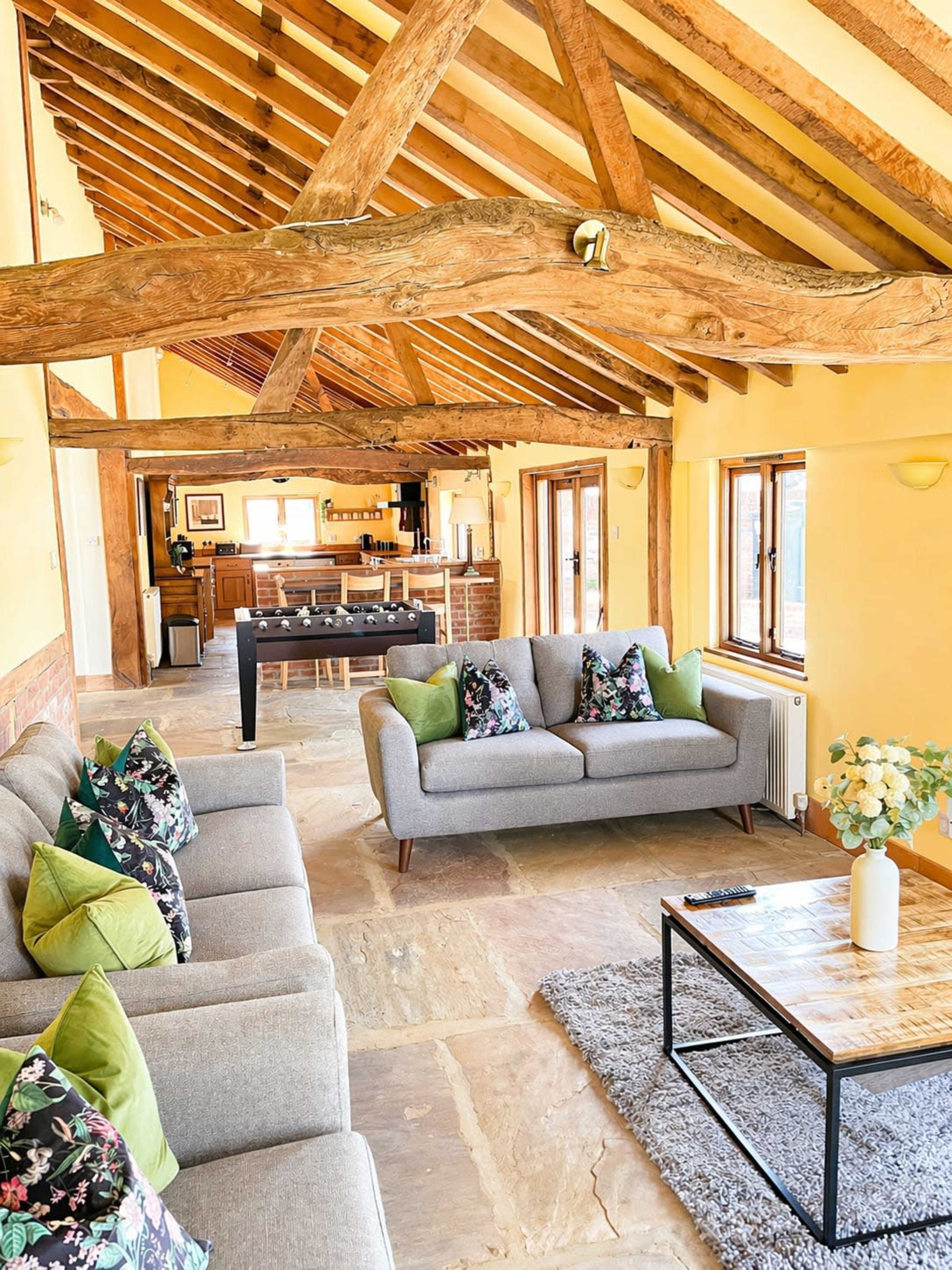 The image shows a spacious living area with wooden beams, natural stone flooring, and two sofas adorned with decorative pillows, facing a wooden coffee table.