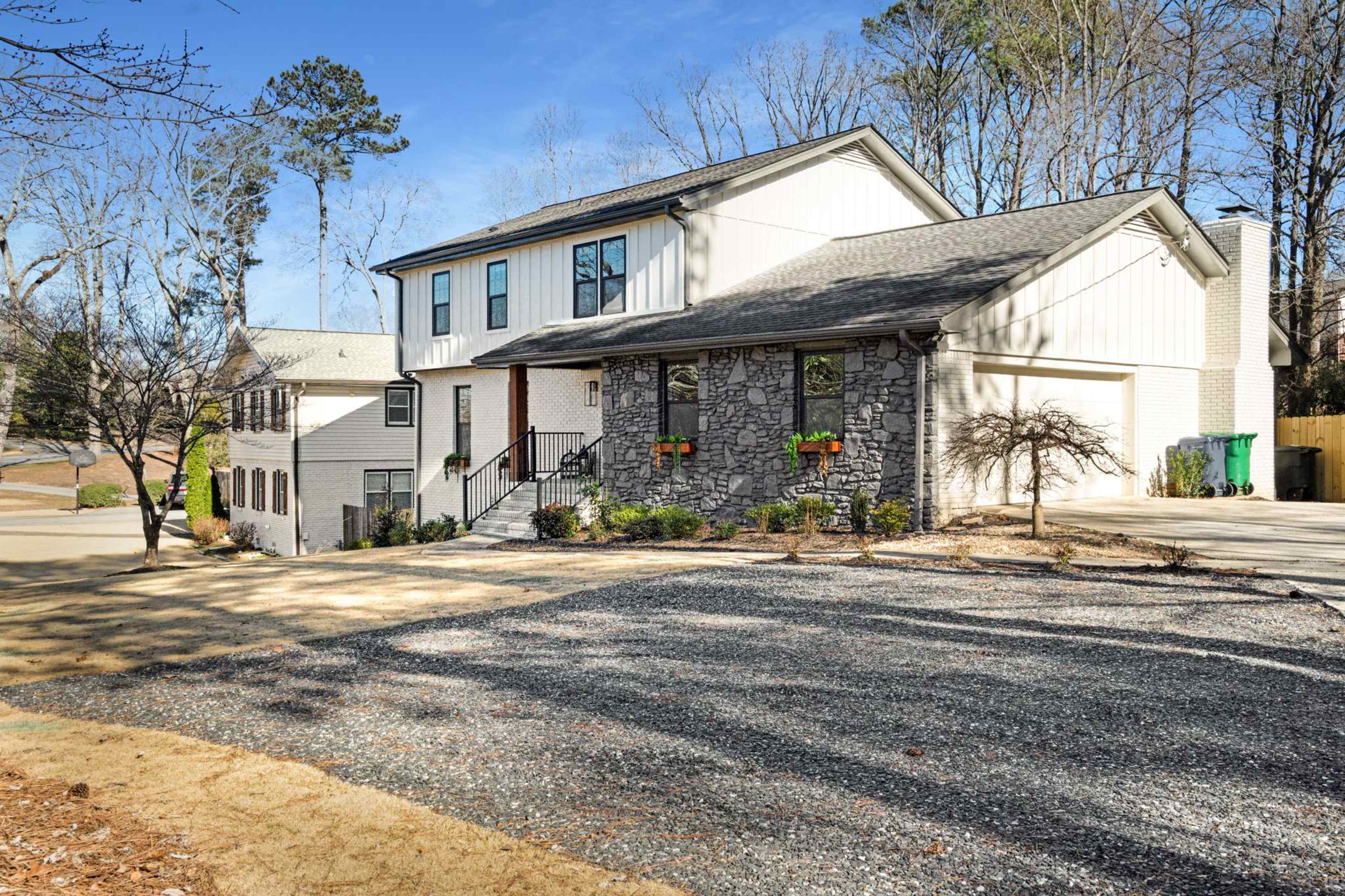 The image shows a two-story house with a stone exterior and large windows, positioned on a gravel driveway surrounded by trees.