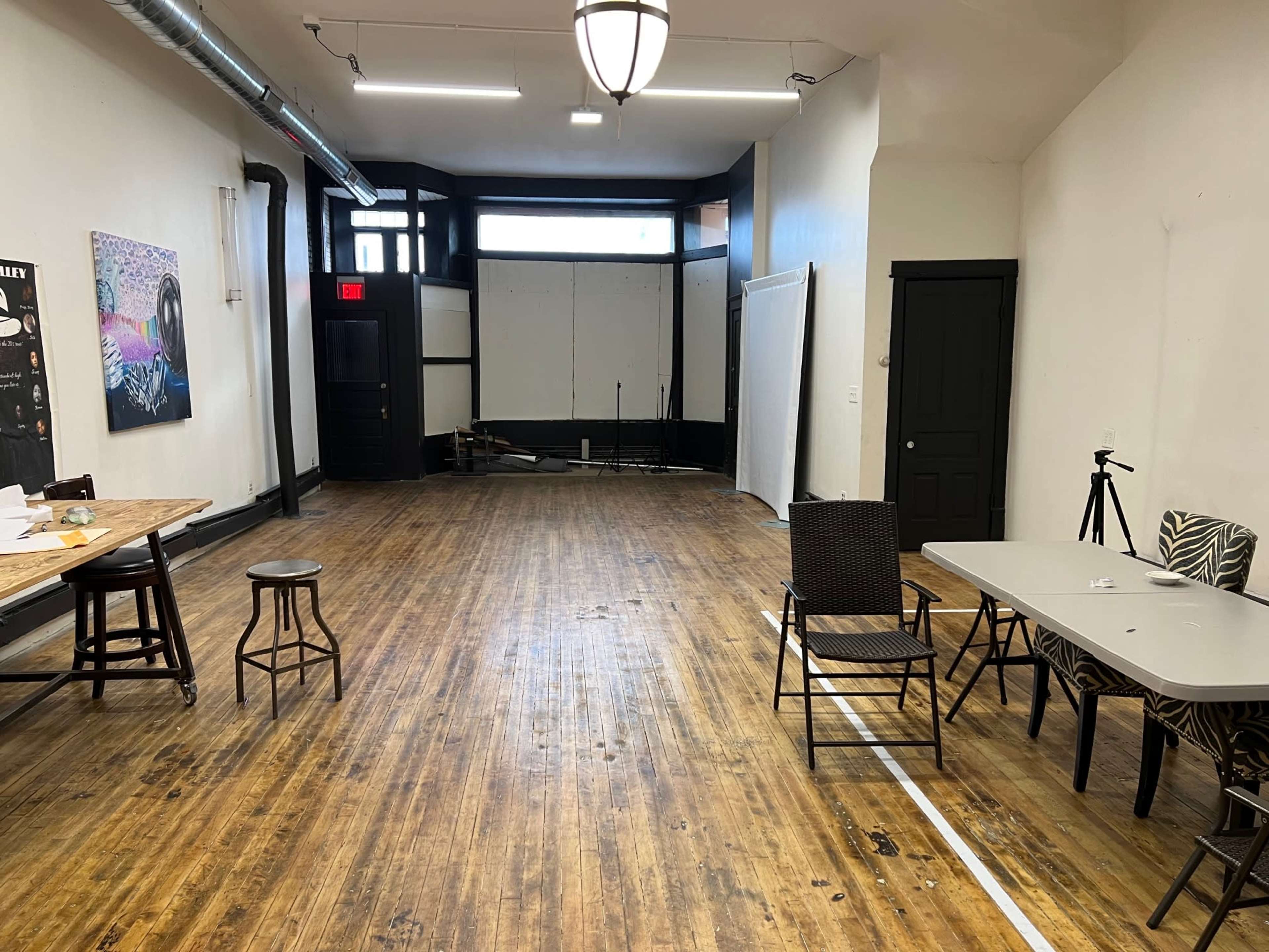 A spacious, empty room with wooden floors, a few tables and chairs, and large windows providing natural light.