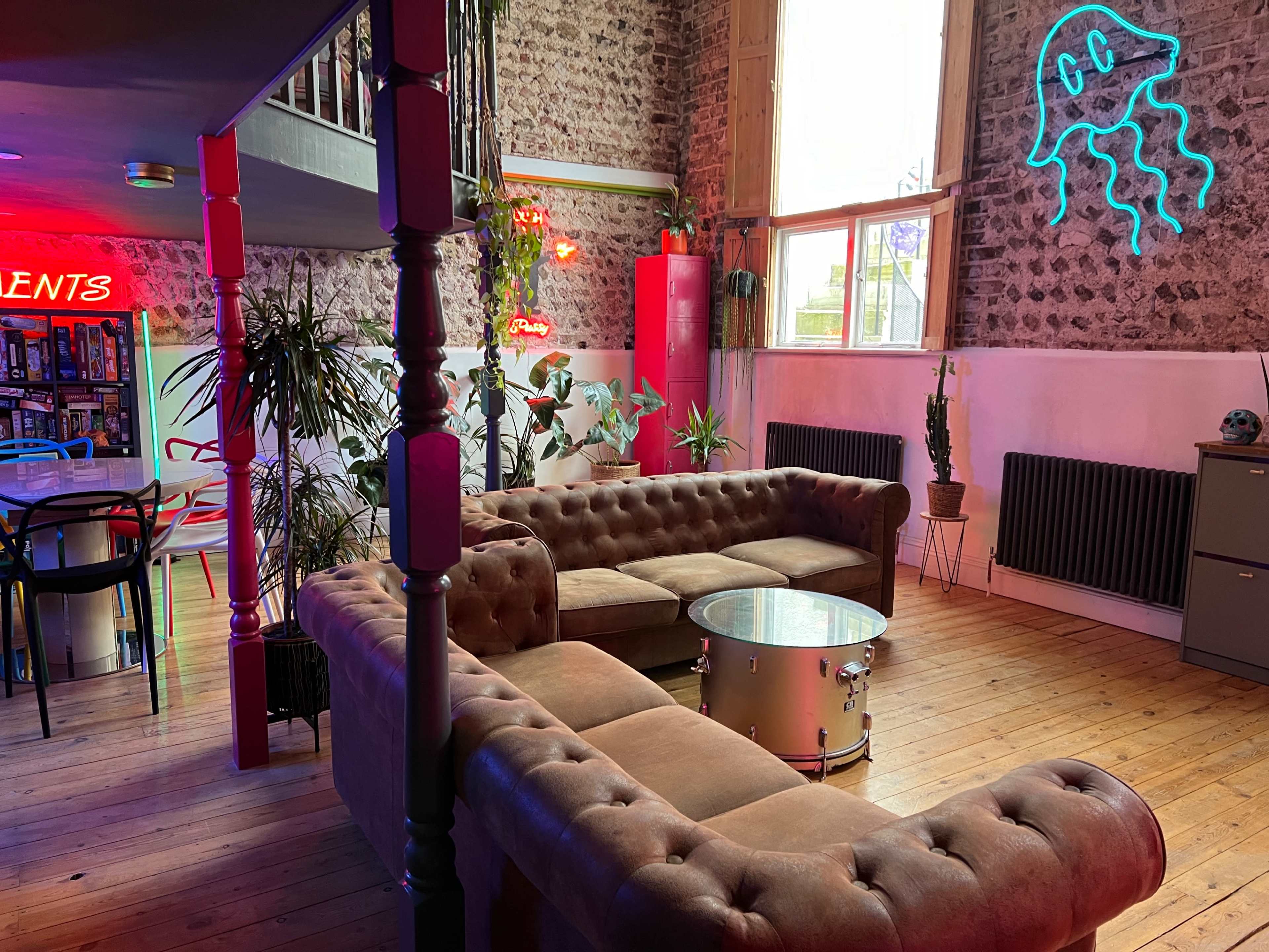 A cozy lounge area with a brown sectional couch, a round table, plants, and neon signs, all set in a room with stone walls and wooden flooring.