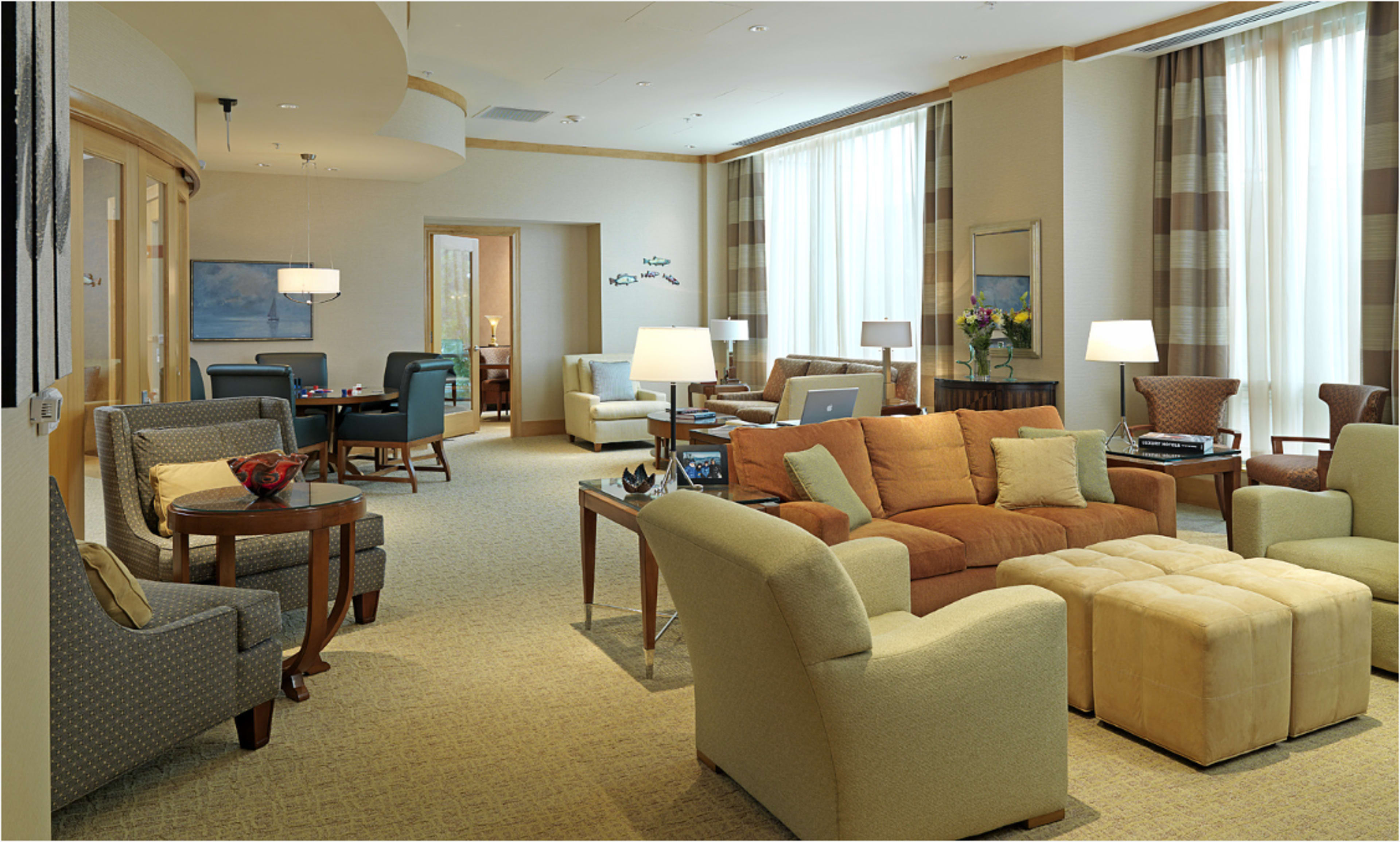 The image shows a spacious and well-furnished living area with a mix of seating options, a dining table in the background, and large windows allowing natural light to enter.