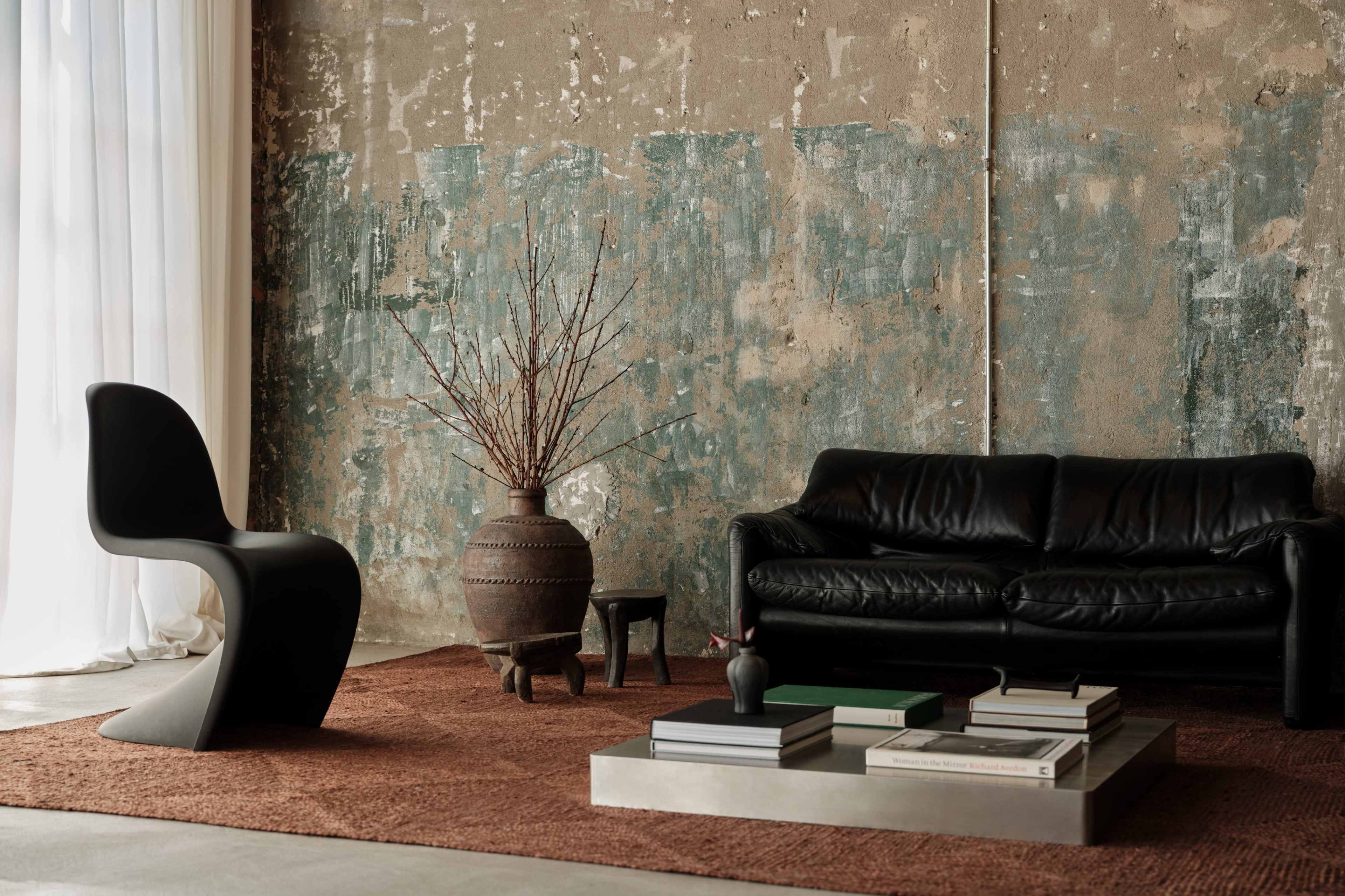 The image features a modern living room with a black leather sofa, a minimalist chair, a coffee table with books, and decorative items set against a textured, weathered wall.