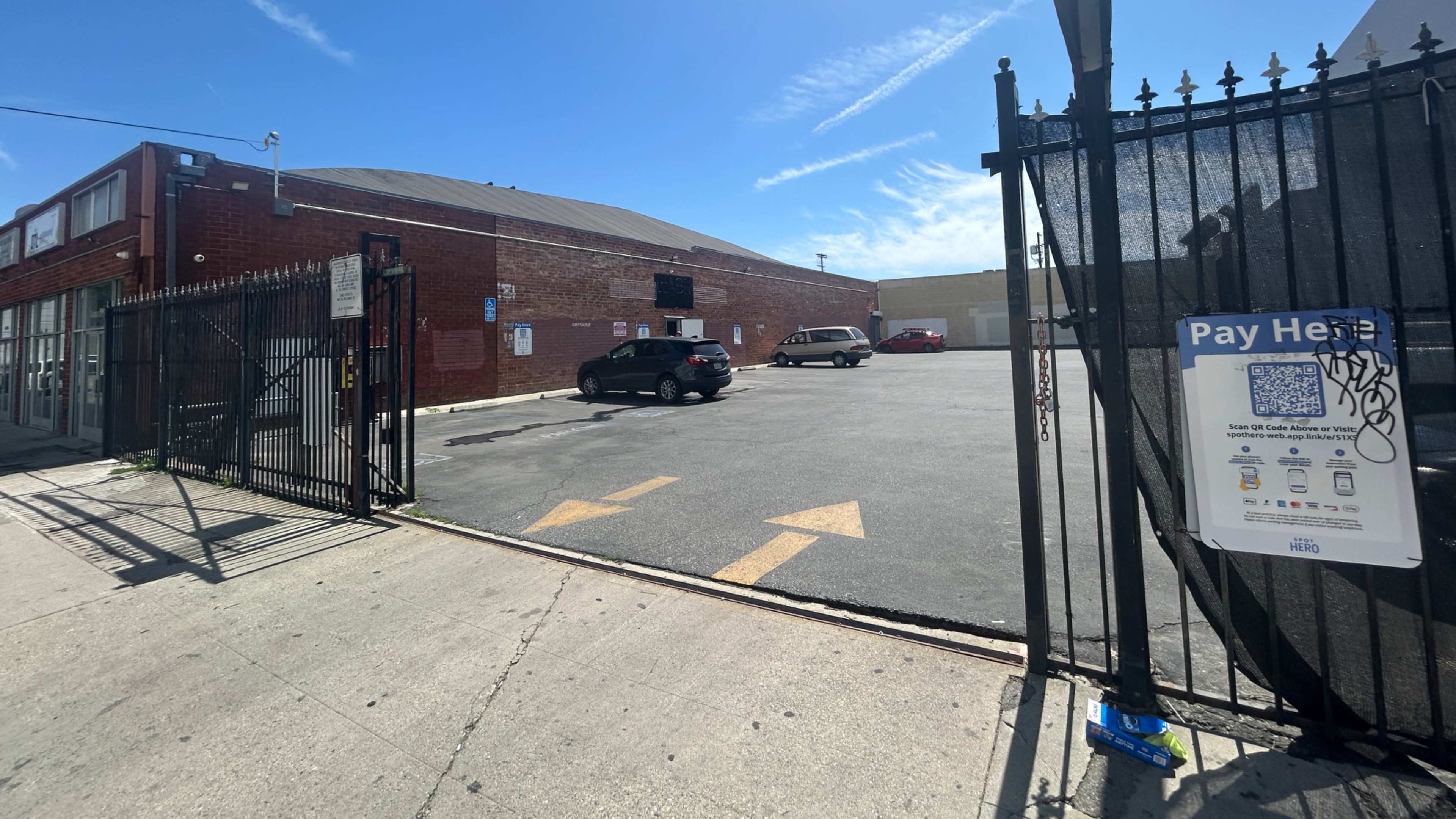 The image shows an empty parking lot enclosed by a gated fence, with a few parked cars and a payment sign visible.