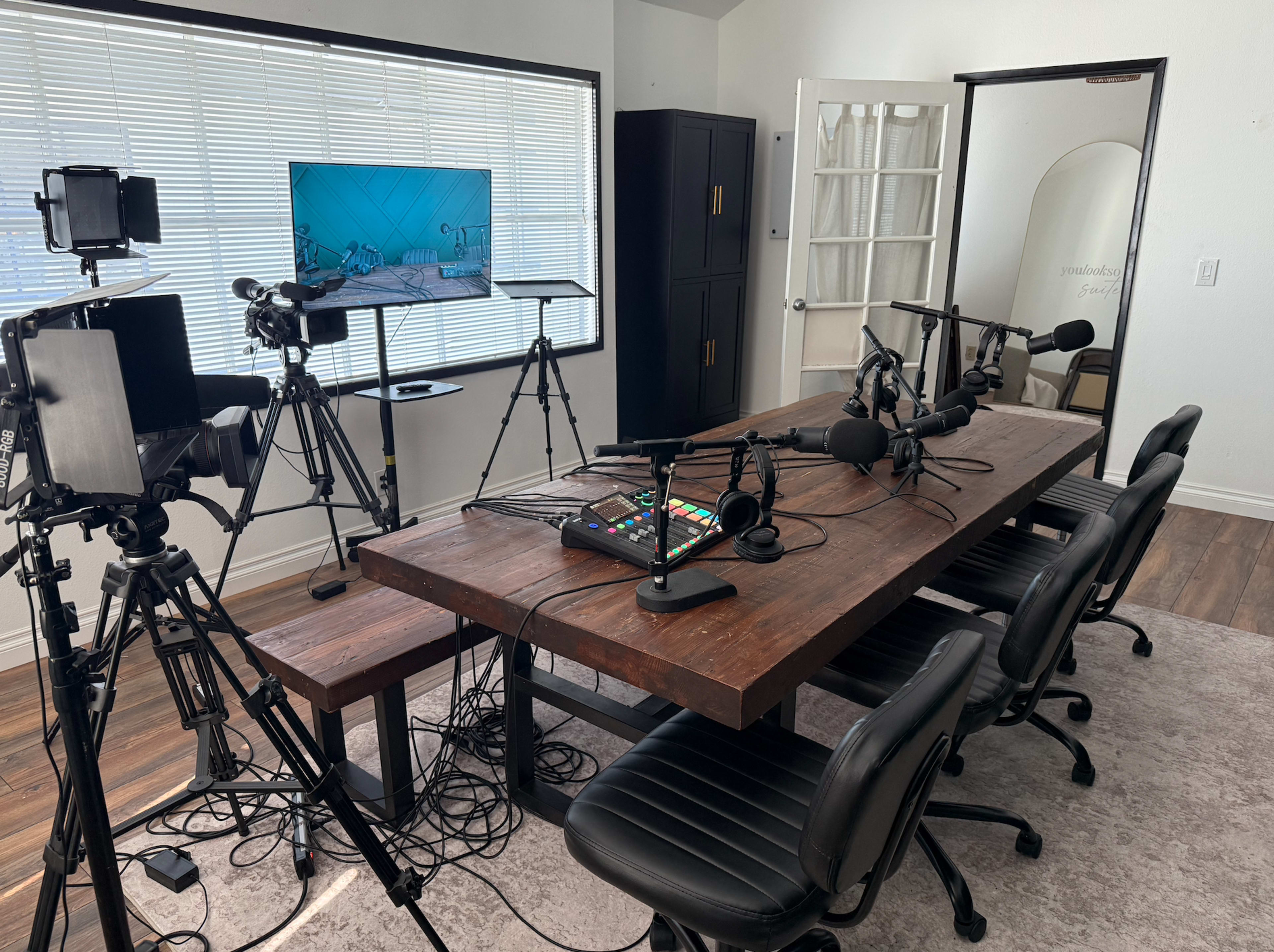 The image shows a recording studio setup with a wooden table surrounded by black chairs, microphones, and cameras, along with a monitor displaying a graphics backdrop.
