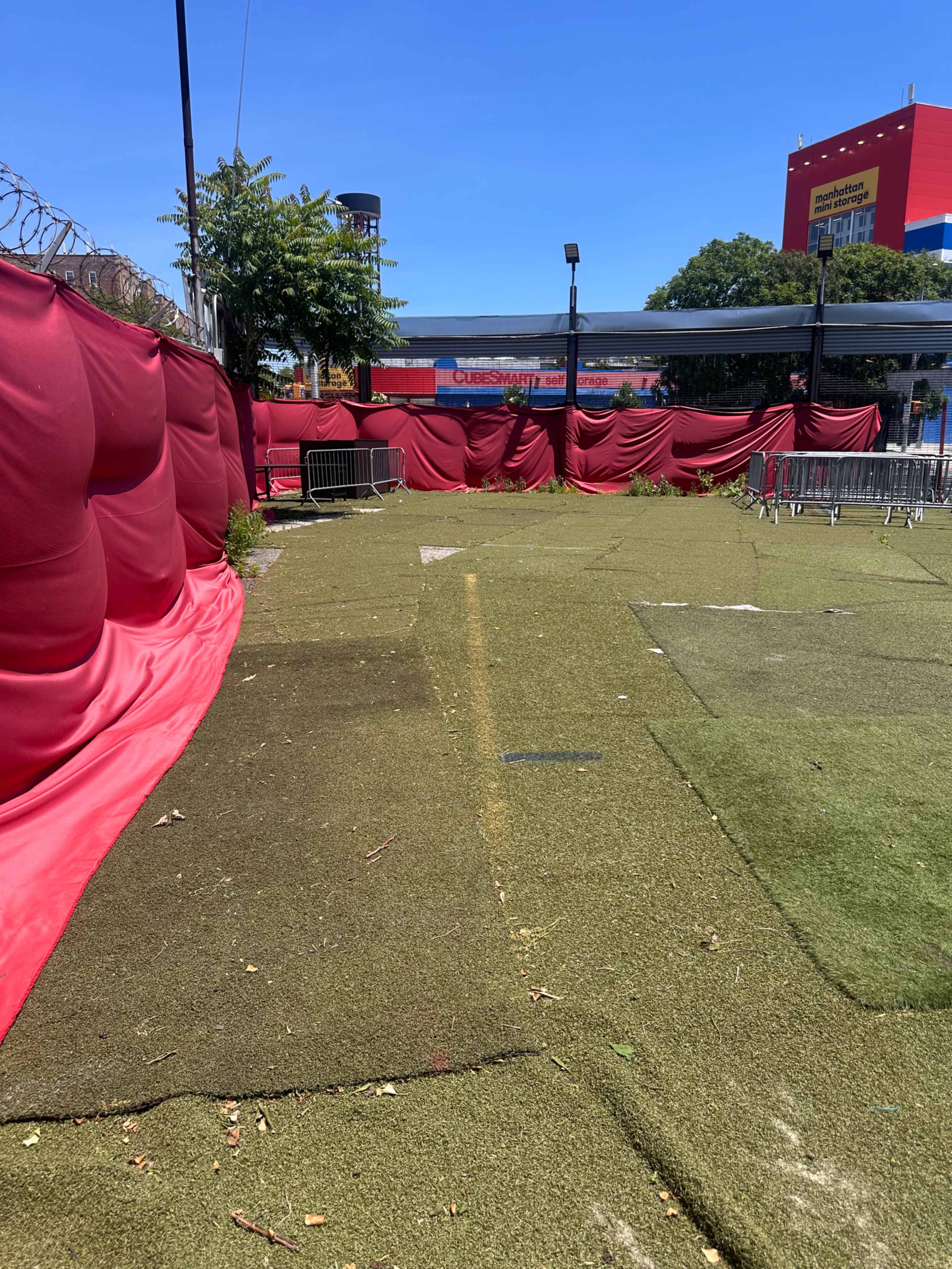 The image shows a fenced area with artificial turf, bordered by red fabric draping on one side and seating barriers scattered throughout.
