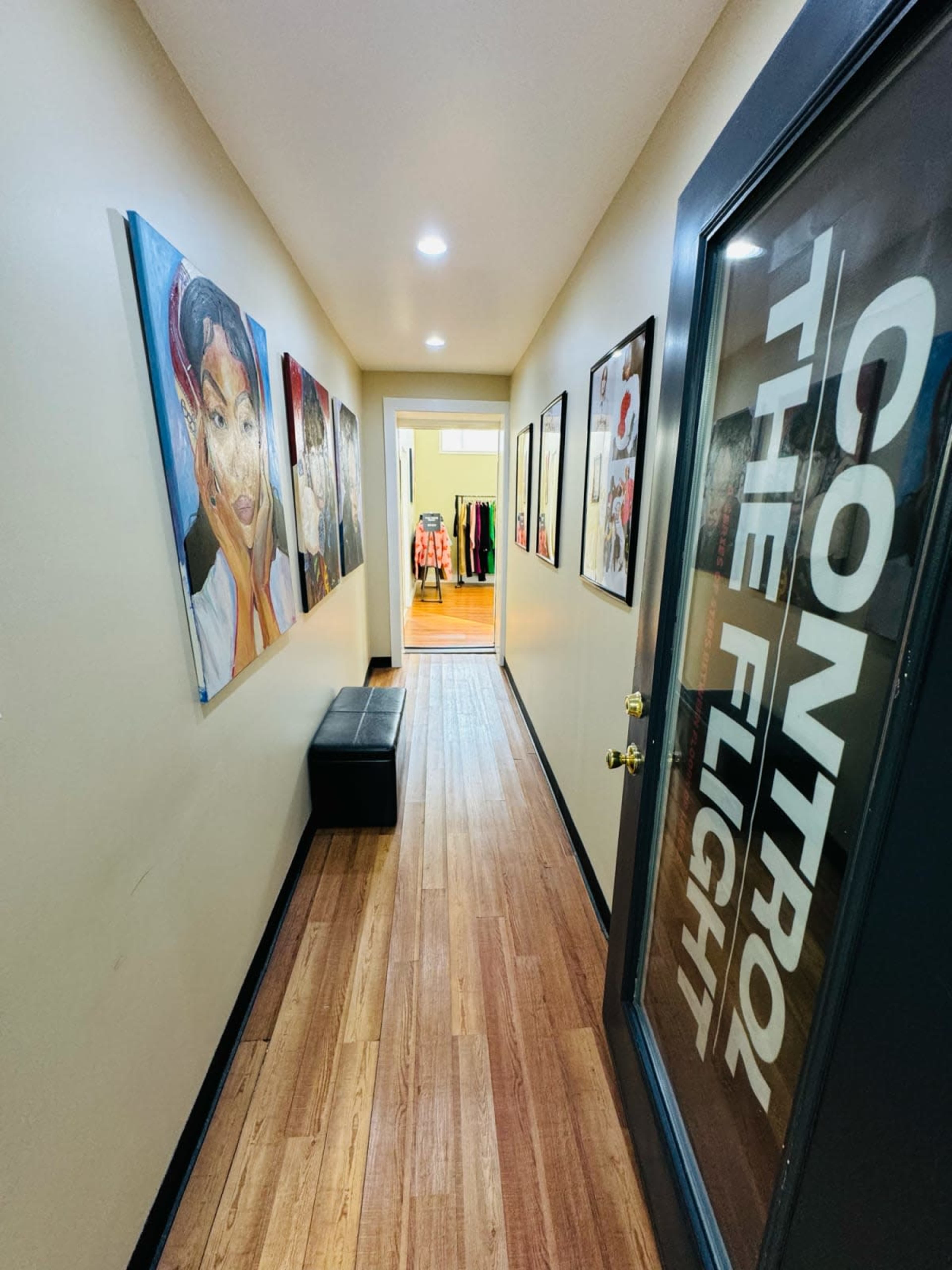The image shows a narrow hallway featuring several framed artworks on the walls, with a black bench and a door at the end leading to another room illuminated by overhead lights.