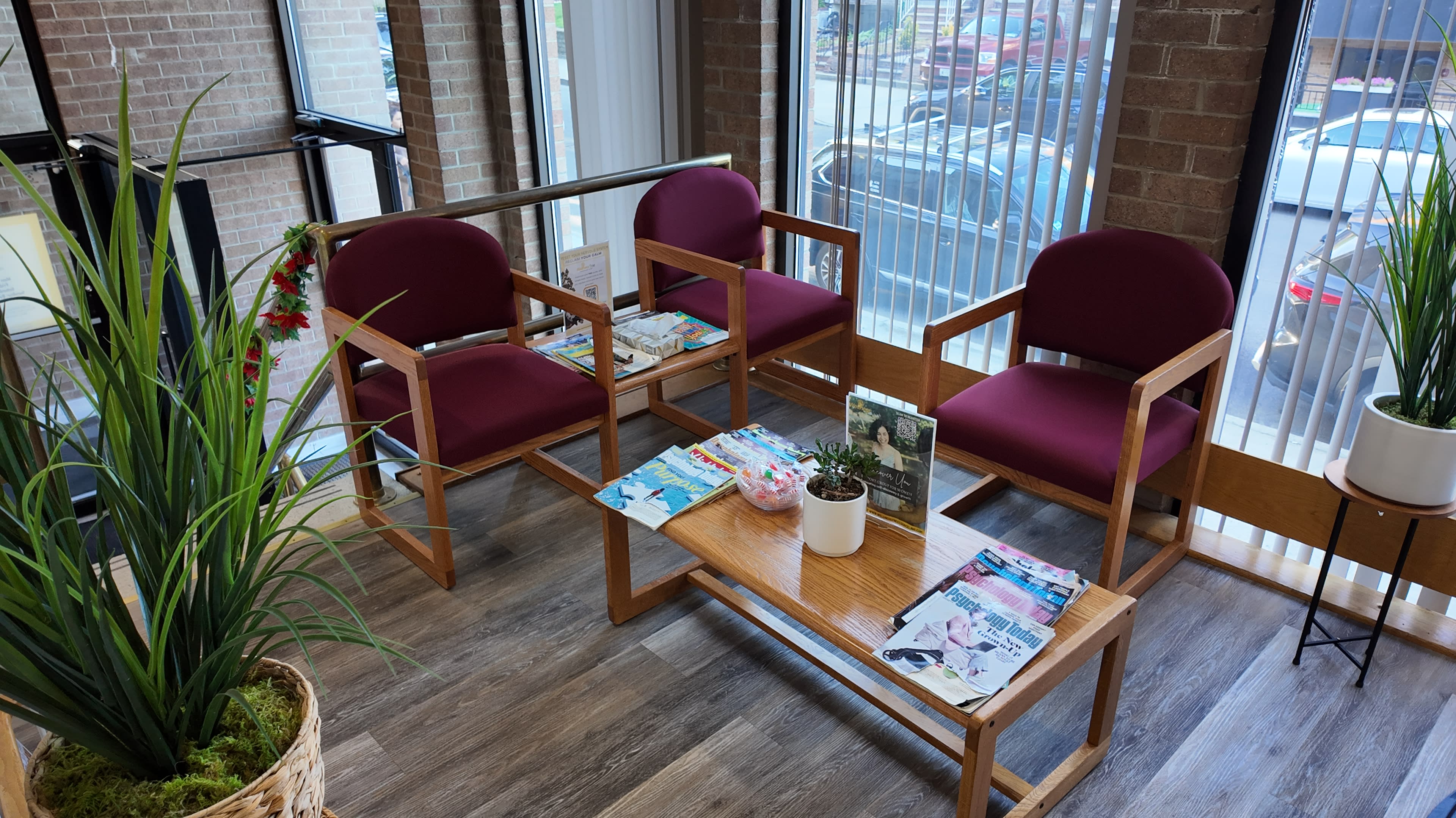 The image shows a waiting area with three maroon chairs and a wooden table, surrounded by potted plants and magazines on the table.