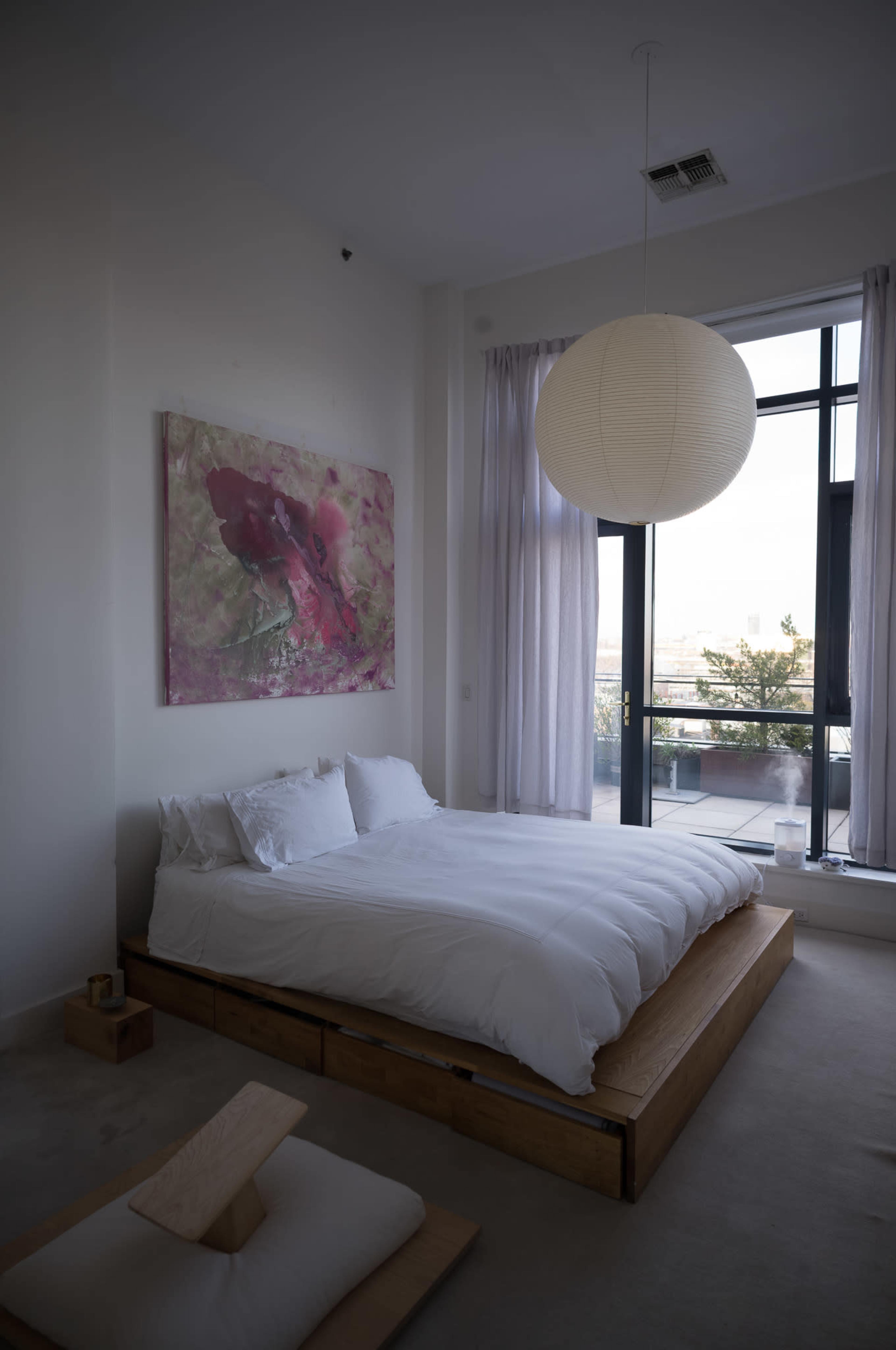 The image shows a minimalist bedroom with a low wooden platform bed, white bedding, a large round paper pendant light, and a window with sheer curtains.