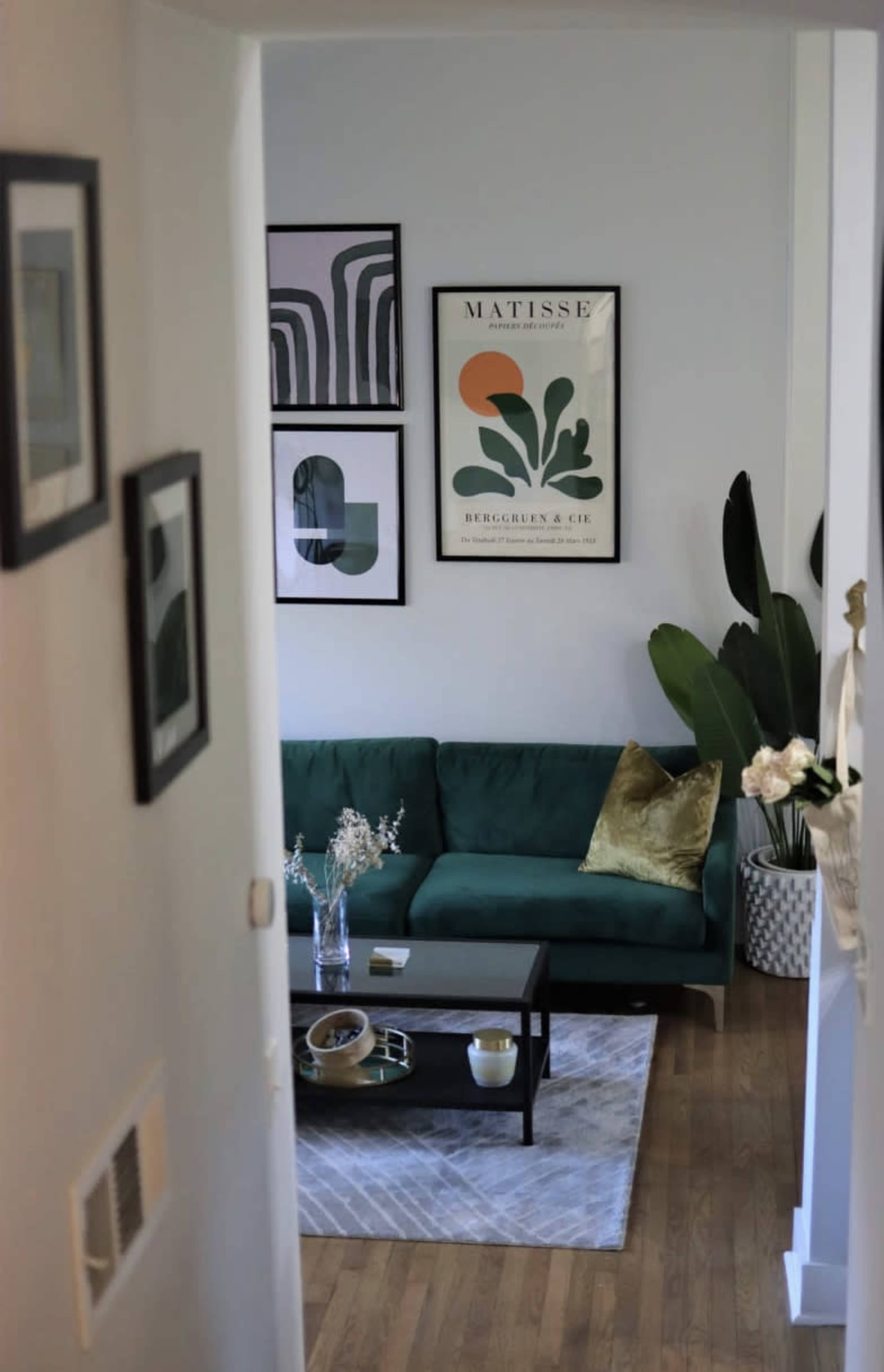 A modern living room features a green sofa, a black coffee table, framed artwork on the walls, and potted plants.