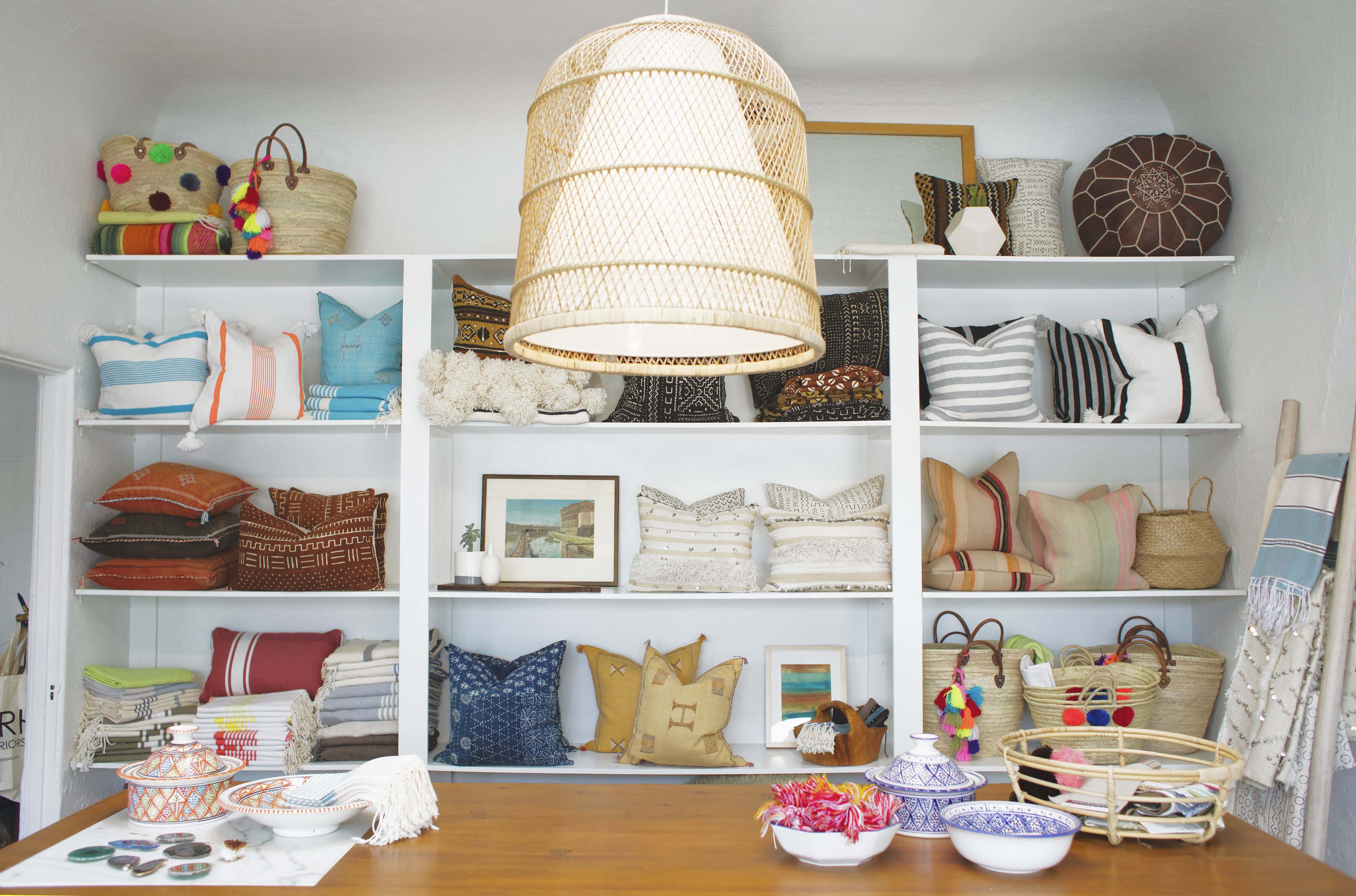 A display of various patterned pillows and woven baskets is arranged on shelves, under a large woven light fixture, in a brightly lit interior space.