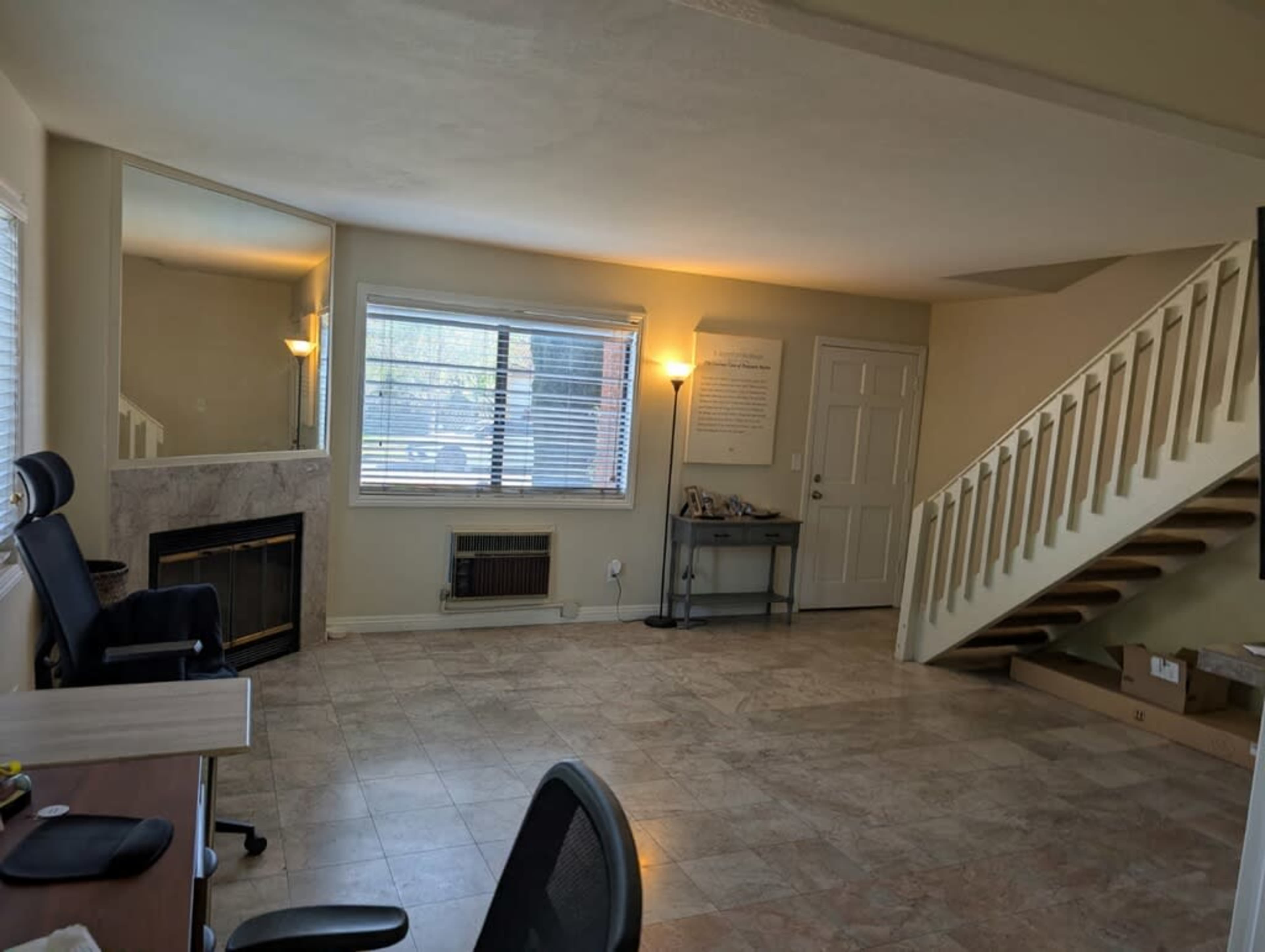 The image shows a spacious living area with a tile floor, a window with blinds, a fireplace, a staircase, and a desk with a black chair.