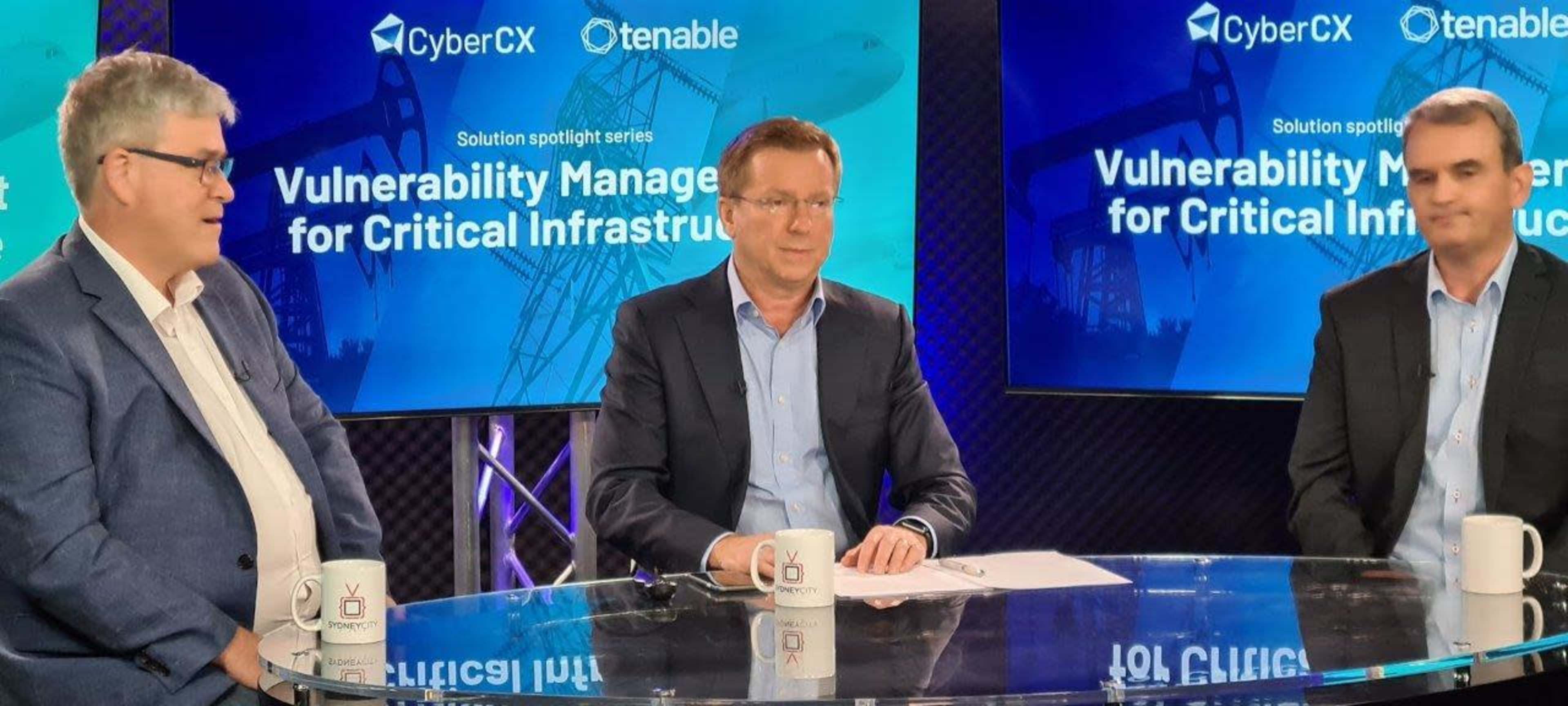 Three professionals sit at a table, discussing vulnerability management for critical infrastructure, with a large screen displaying the topic in the background.