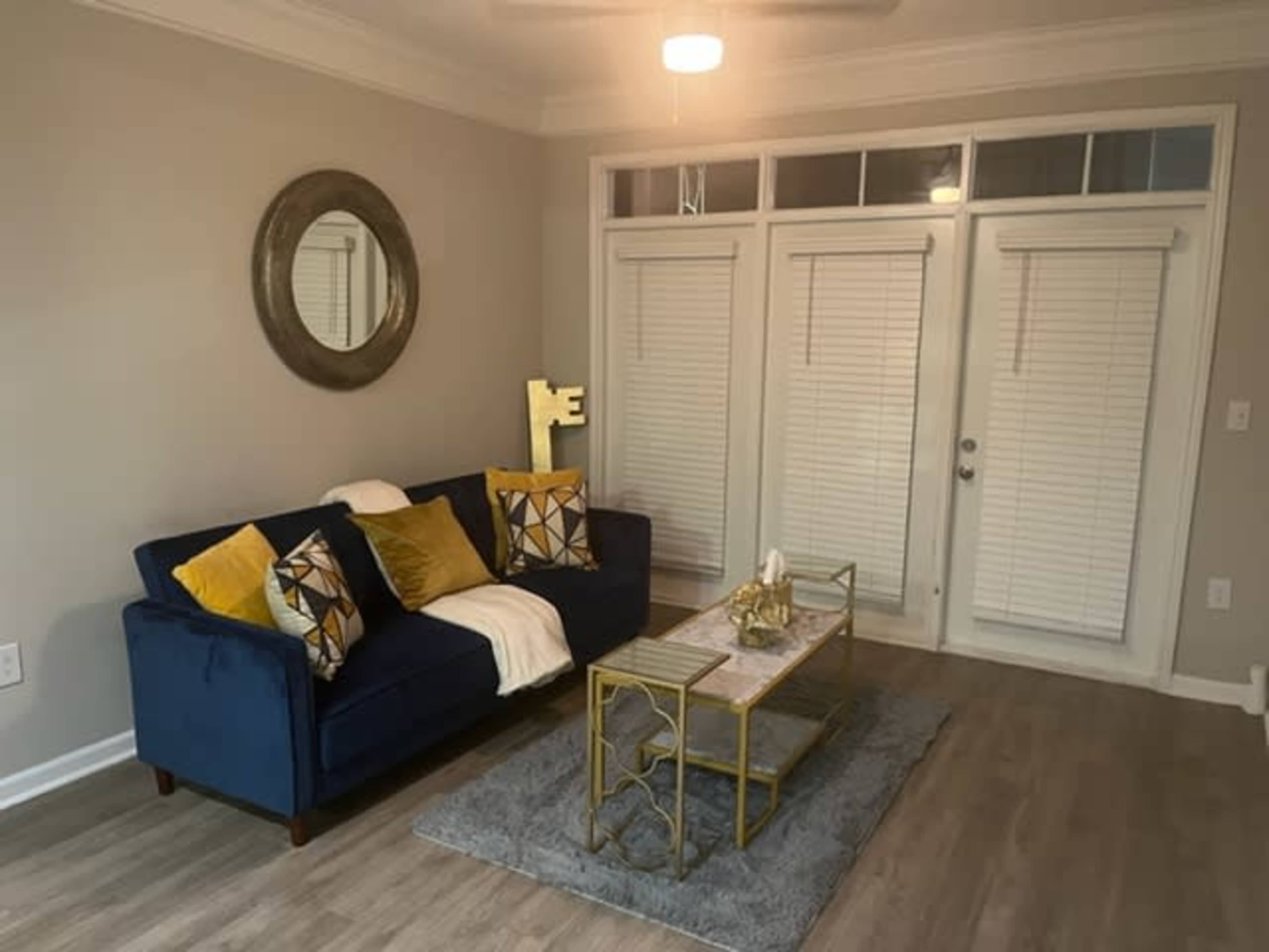 The image showcases a living room featuring a blue sofa with yellow and black patterned cushions, a marble-topped coffee table, and large windows with white blinds.