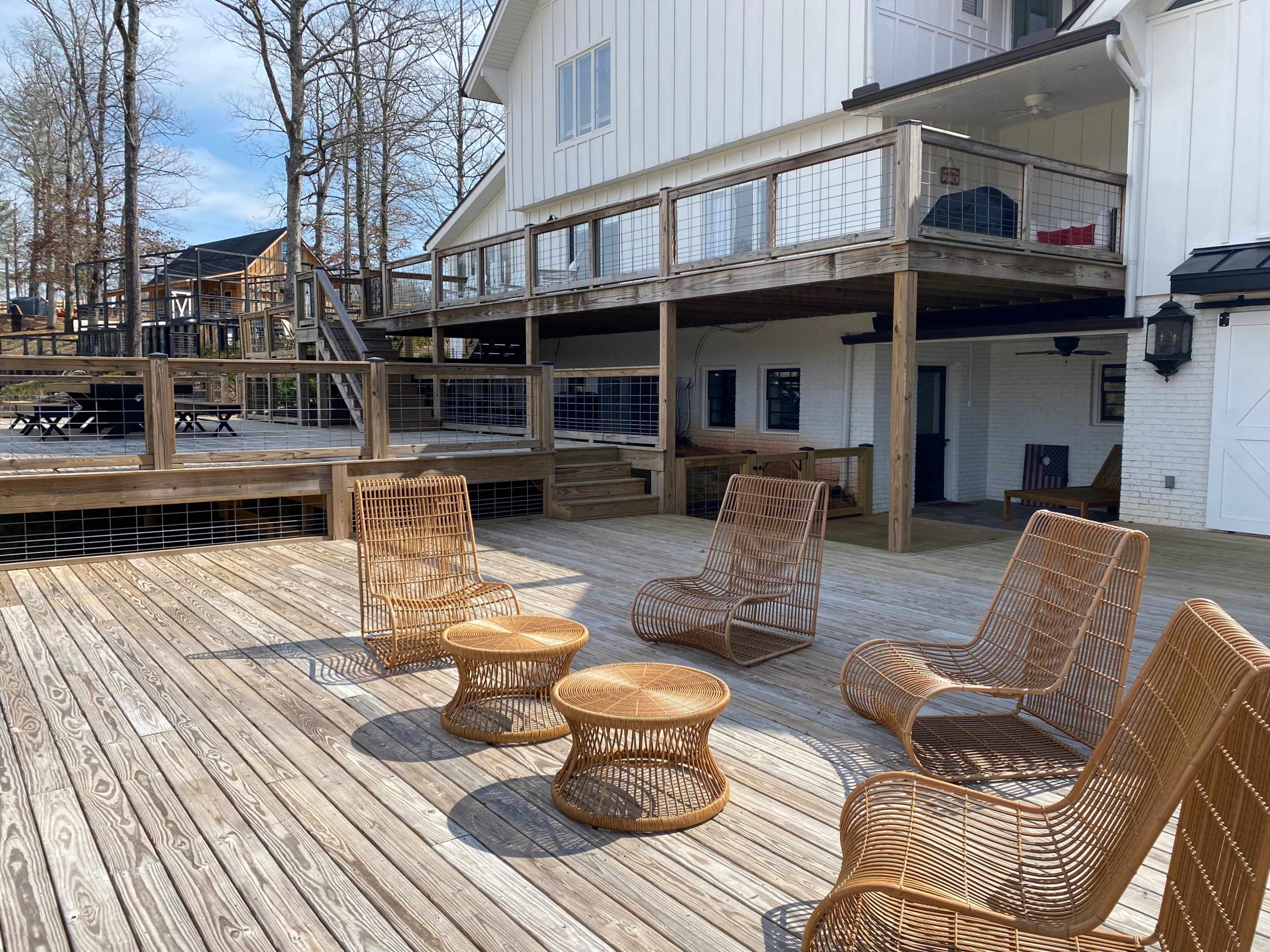The image shows a wooden deck with four rattan chairs and two small tables, adjacent to a white house and surrounded by trees.
