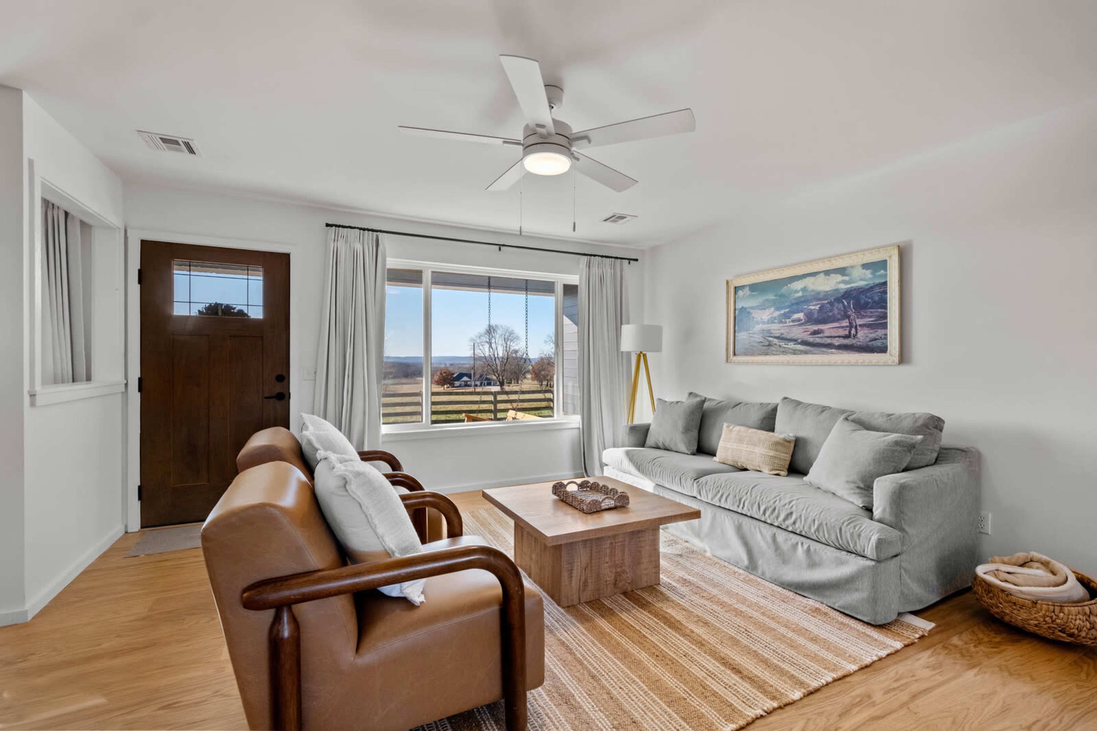 The living room features a light-colored sofa, two brown leather chairs, and a wooden coffee table, with large windows providing a view of the outdoors.