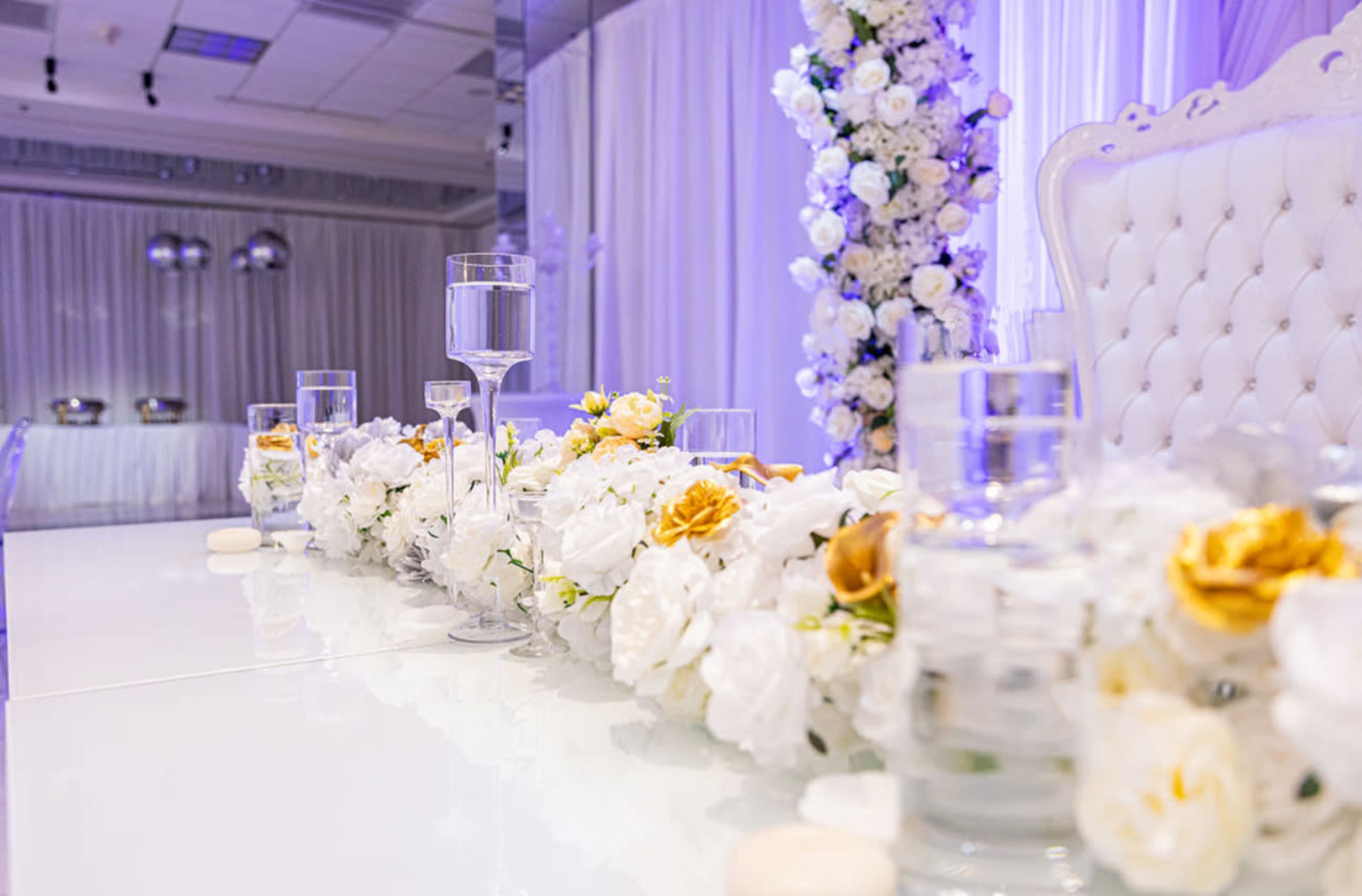 A beautifully arranged table features a floral centerpiece with white and gold flowers, surrounded by clear glasses and elegant decor in a softly lit event space.