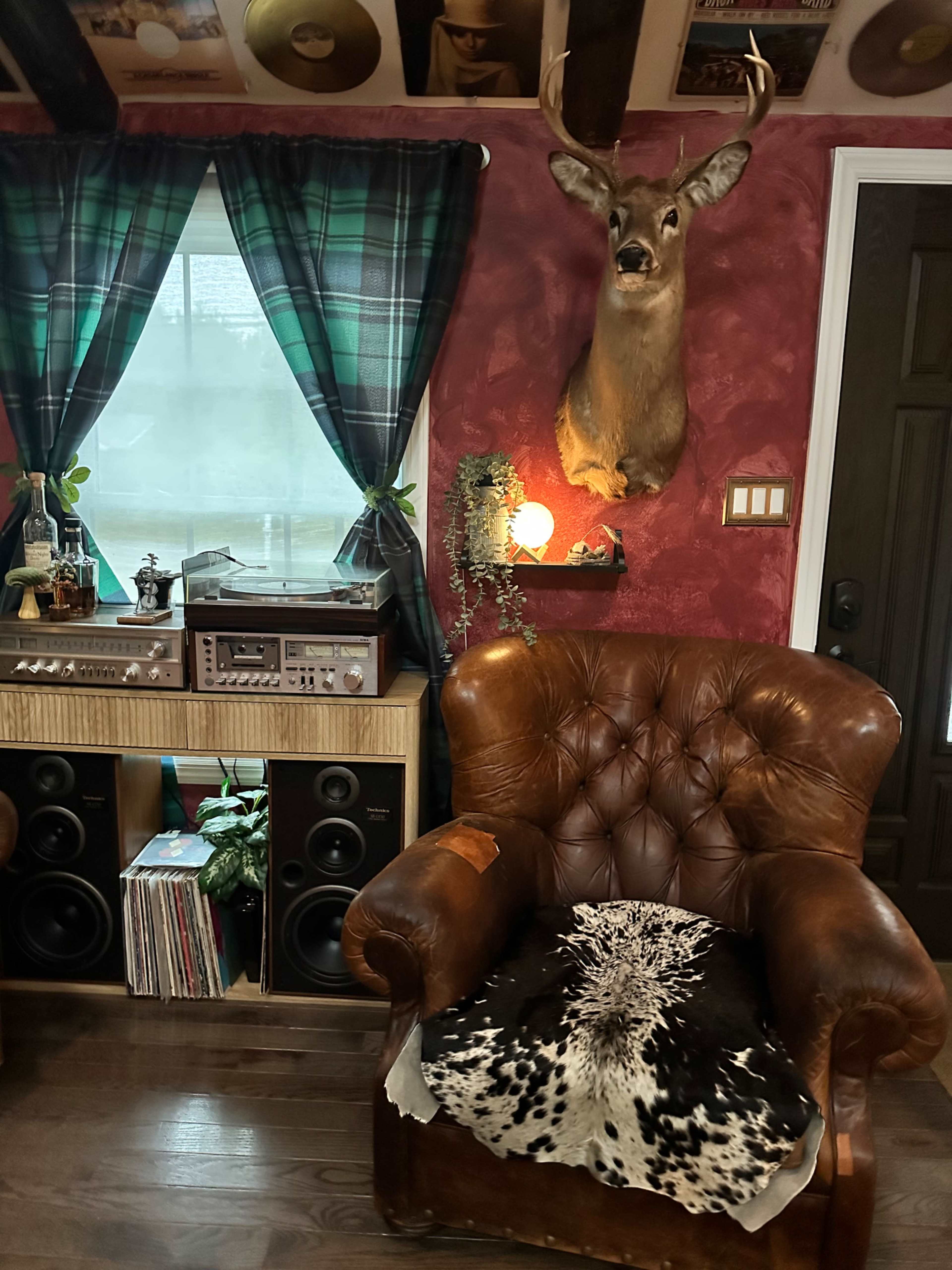 The image shows a cozy room featuring a brown leather armchair, a mounted deer head on the wall, green checkered curtains, a stereo system with speakers, and a collection of records.