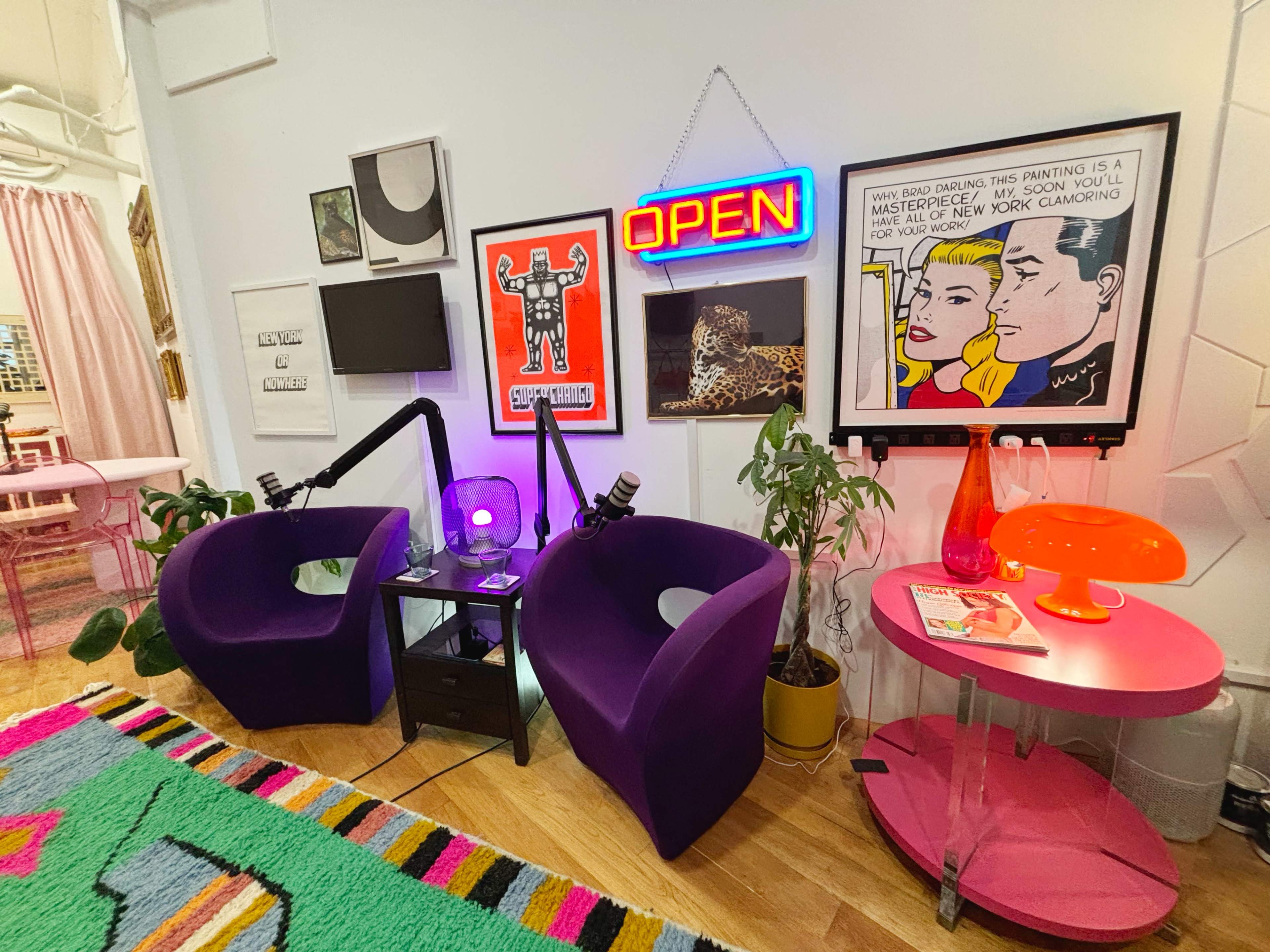 The image shows a colorful lounge area with two purple chairs, a neon "OPEN" sign, artwork on the walls, and a vibrant rug.