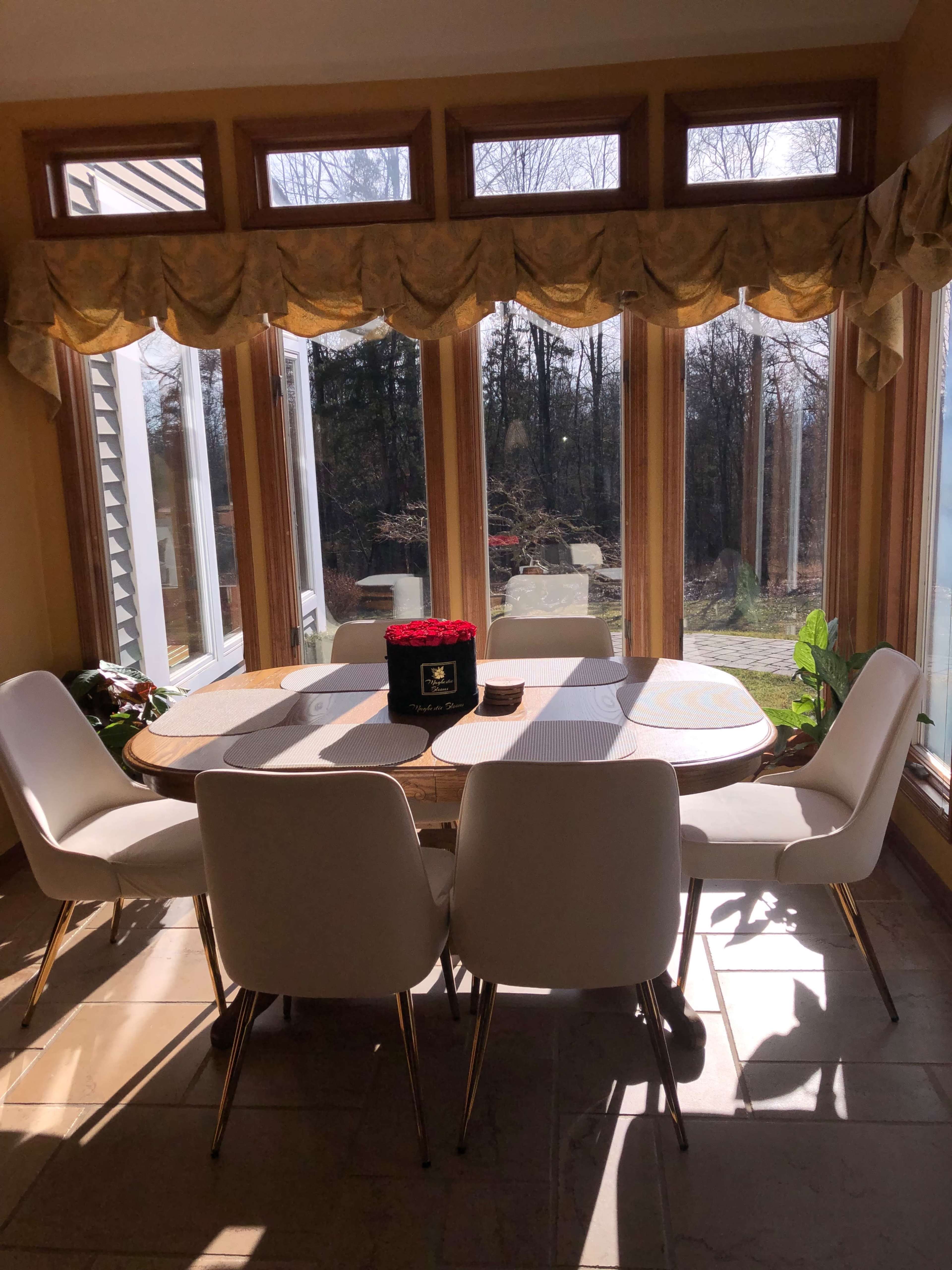A dining table with six white chairs is positioned in a sunlit area with large windows, revealing a view of a garden.