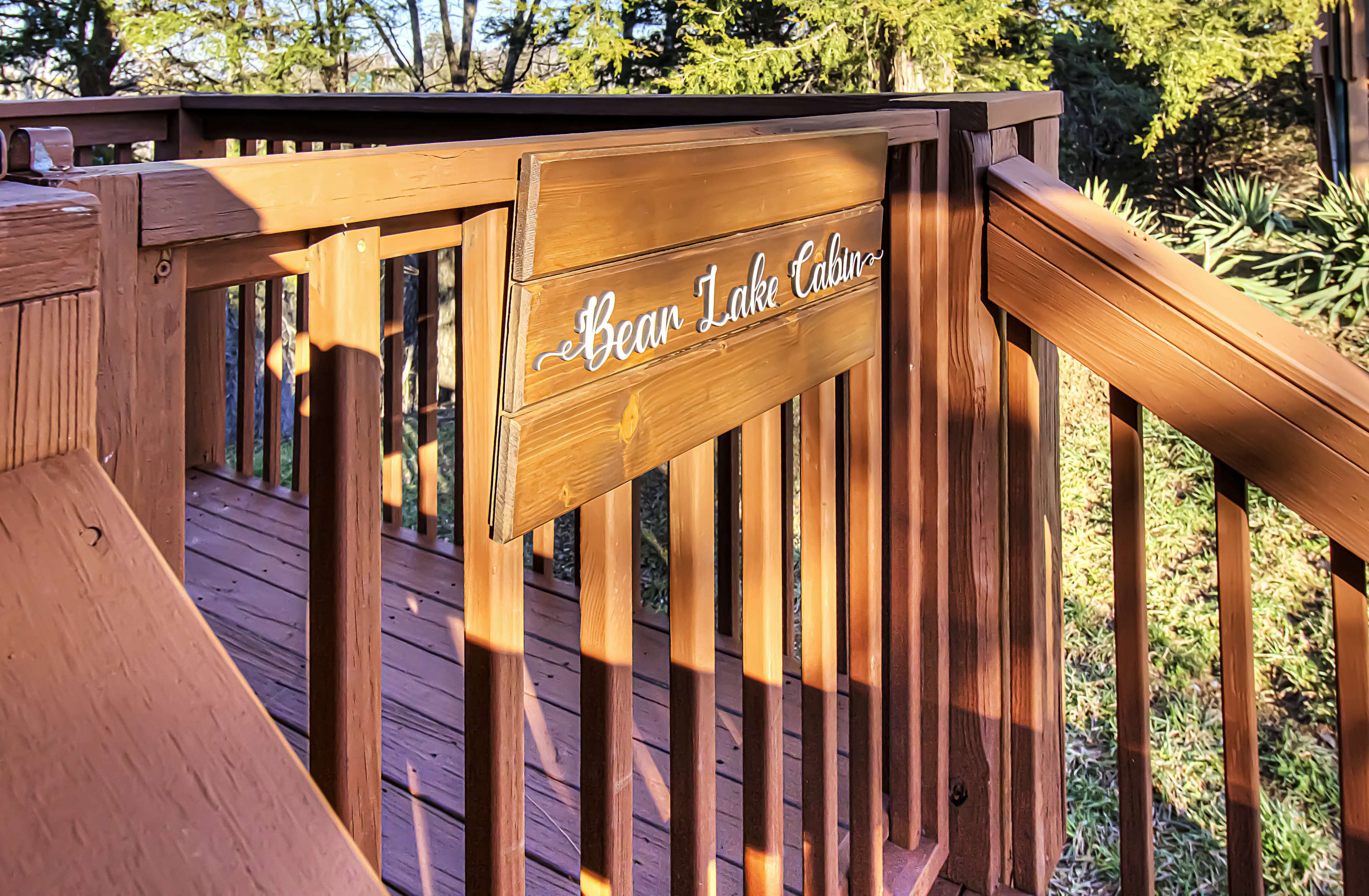 A wooden sign labeled "Bear Lake Cabin" is mounted on a railing leading to a deck surrounded by greenery.