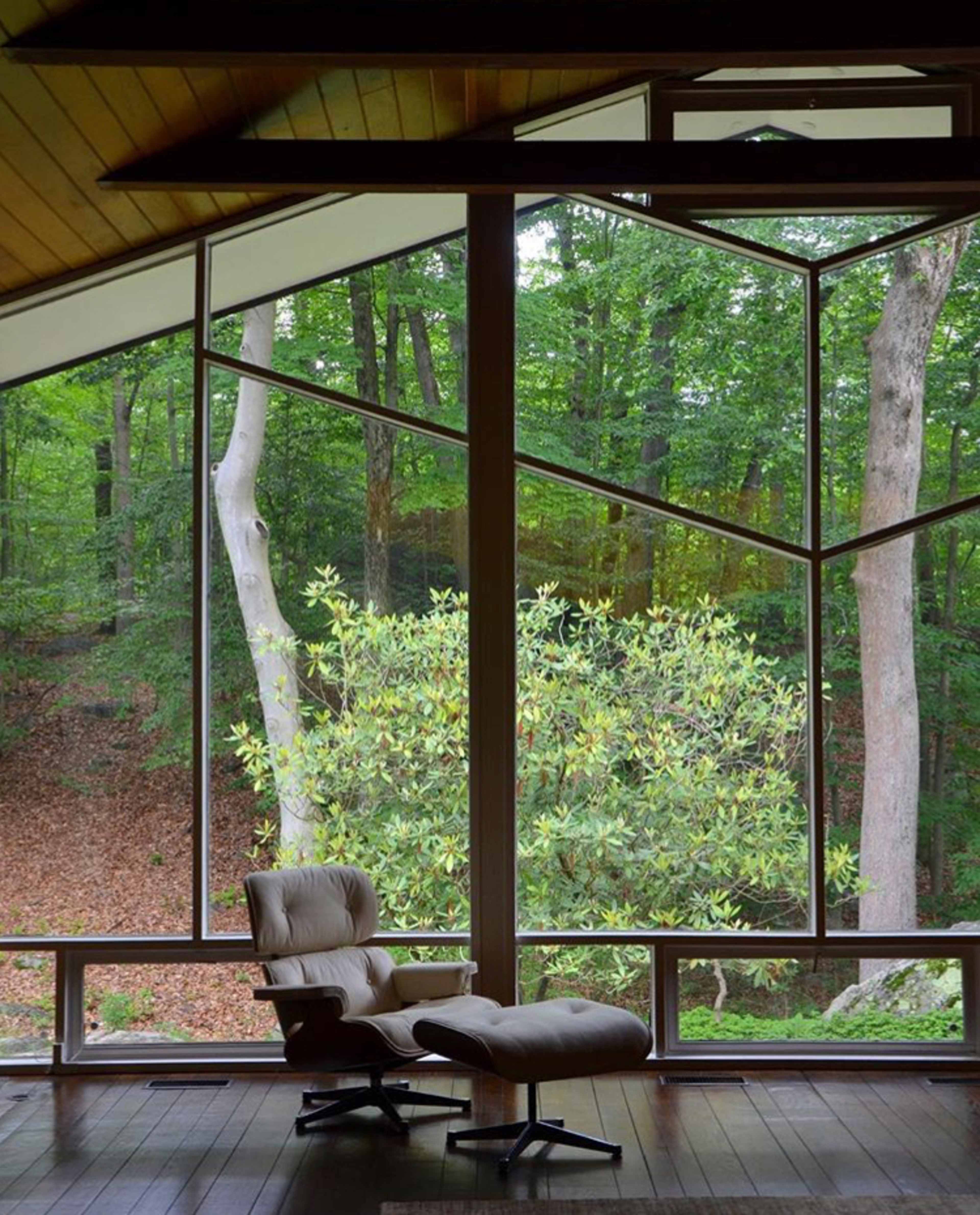 A modern interior featuring a reclined chair beside a large geometric window that offers a view of a wooded landscape.