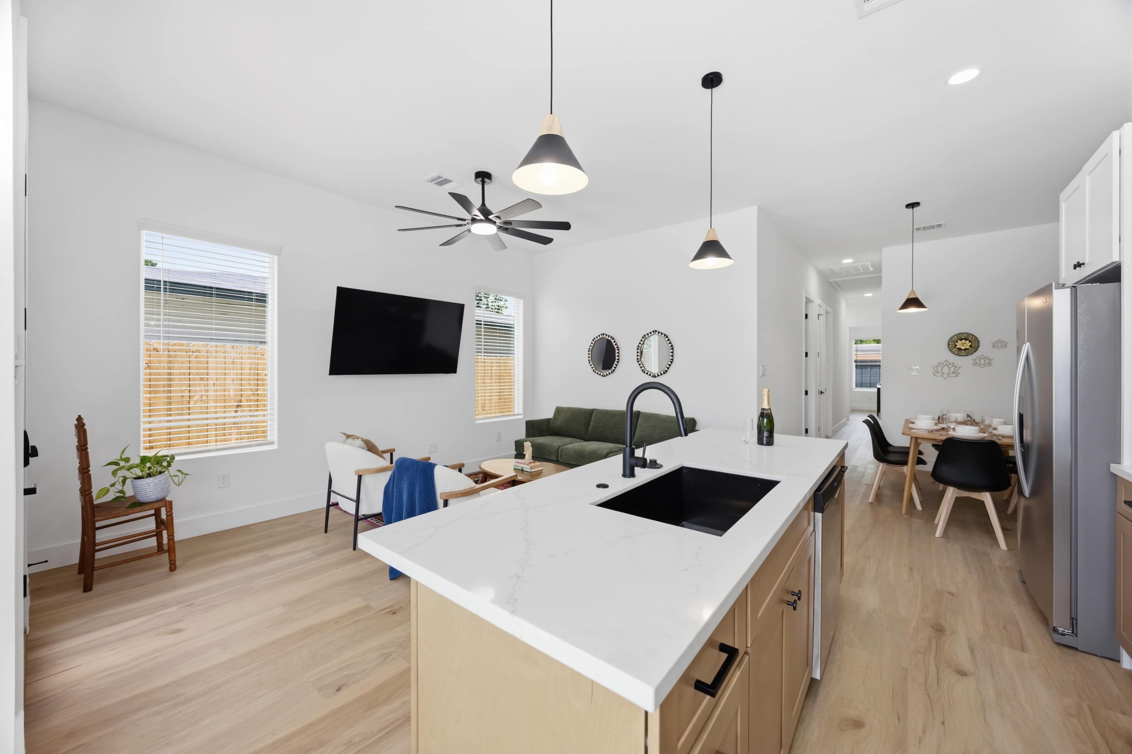 The image shows a modern living space featuring an open layout with a kitchen, dining area, and living room, illuminated by pendant lights and a ceiling fan.