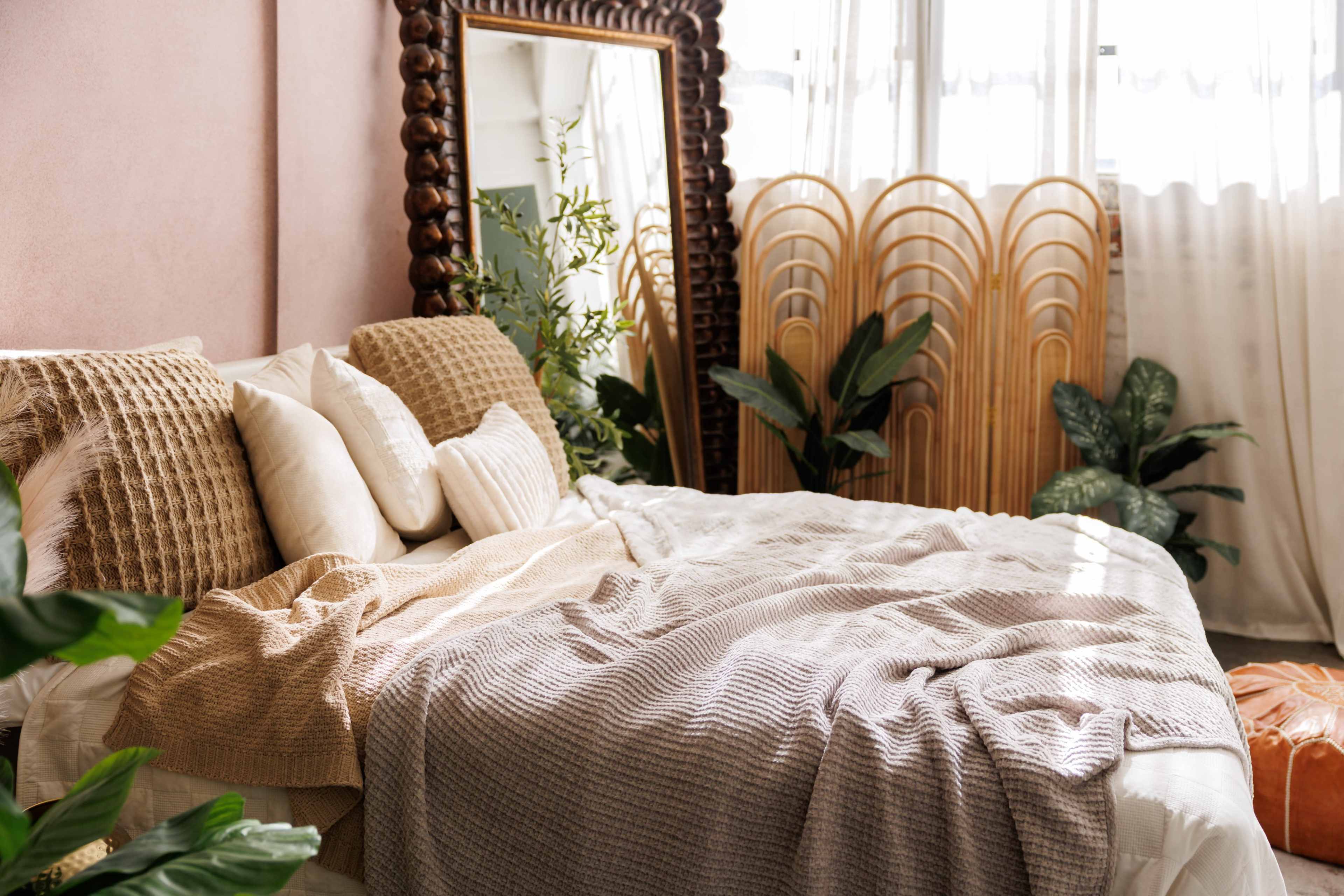 The image shows a cozy bedroom featuring a large bed with various textured pillows and a blanket, surrounded by indoor plants and a decorative mirror.