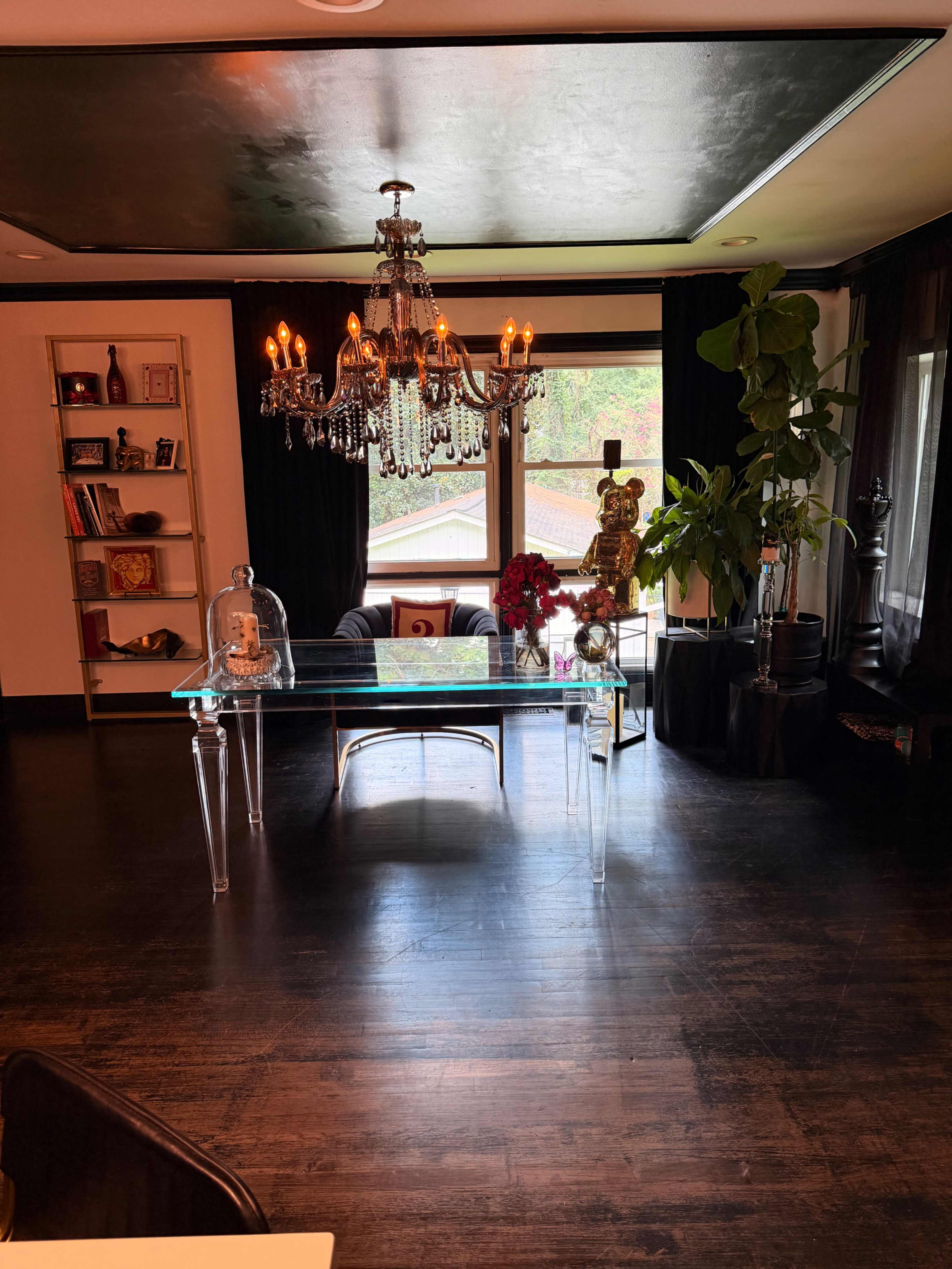 The image shows a stylish room featuring a glass dining table beneath a chandelier, with large windows and dark wood flooring.