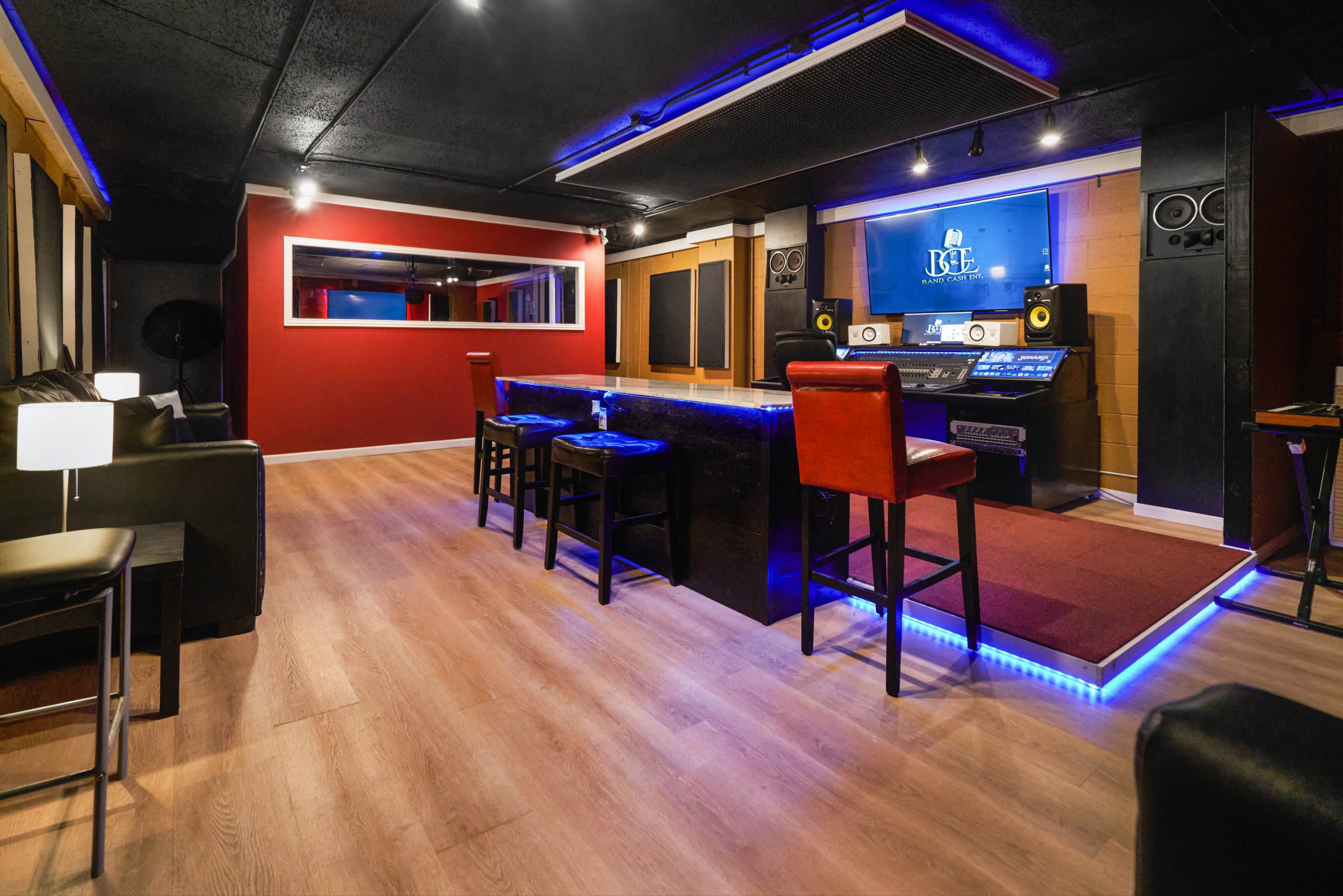 The image shows a recording studio with a central mixing console, bar seating, black and red walls, and acoustic panels.