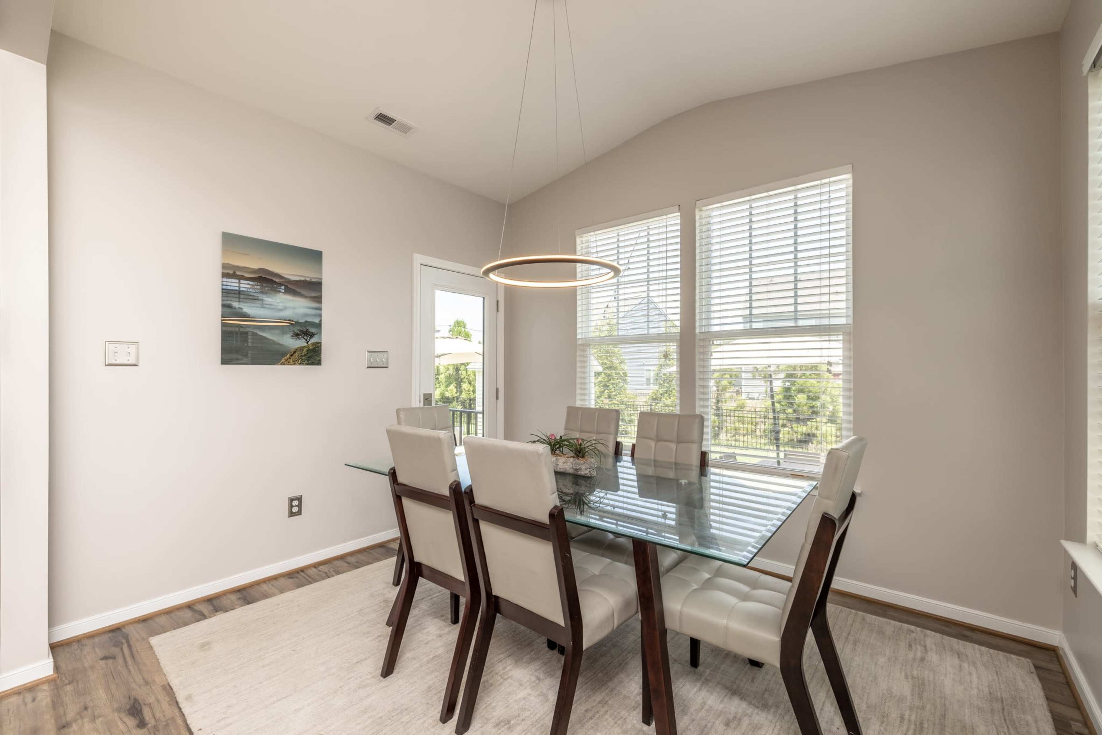A modern dining area features a glass table surrounded by upholstered chairs and a circular pendant light, with large windows allowing natural light to enter.