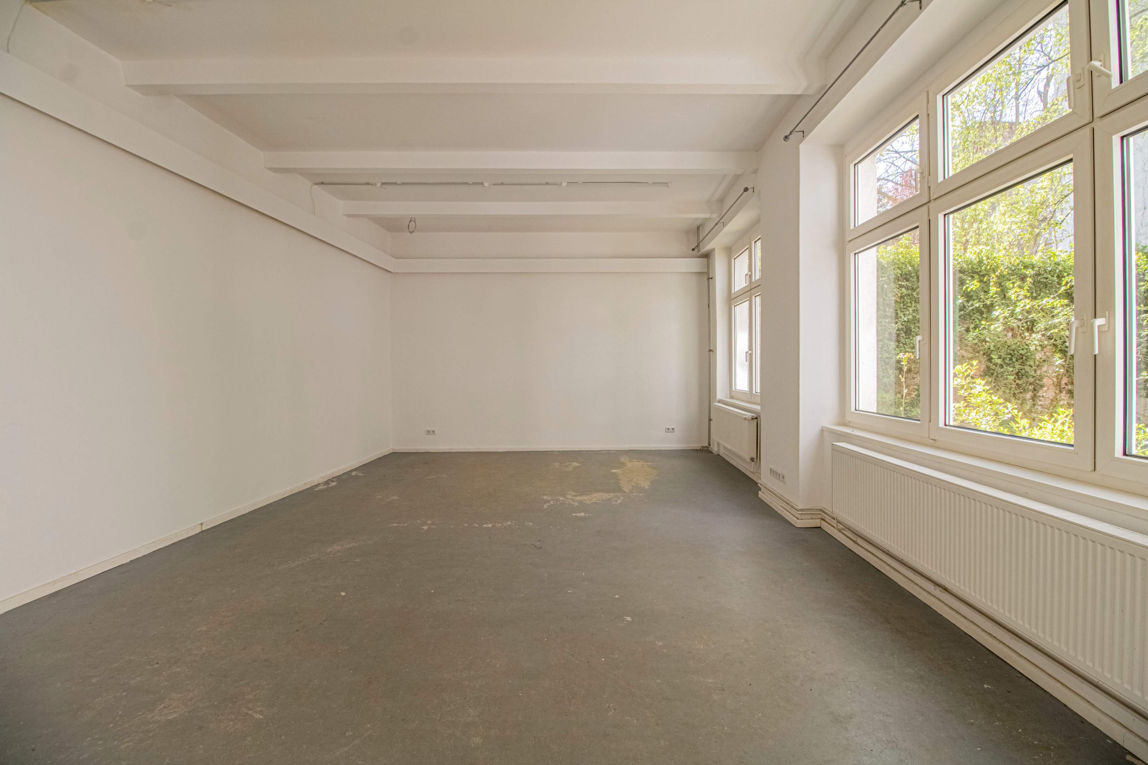 The image shows a vacant room with a plain, light-colored wall and several large windows that let in natural light.