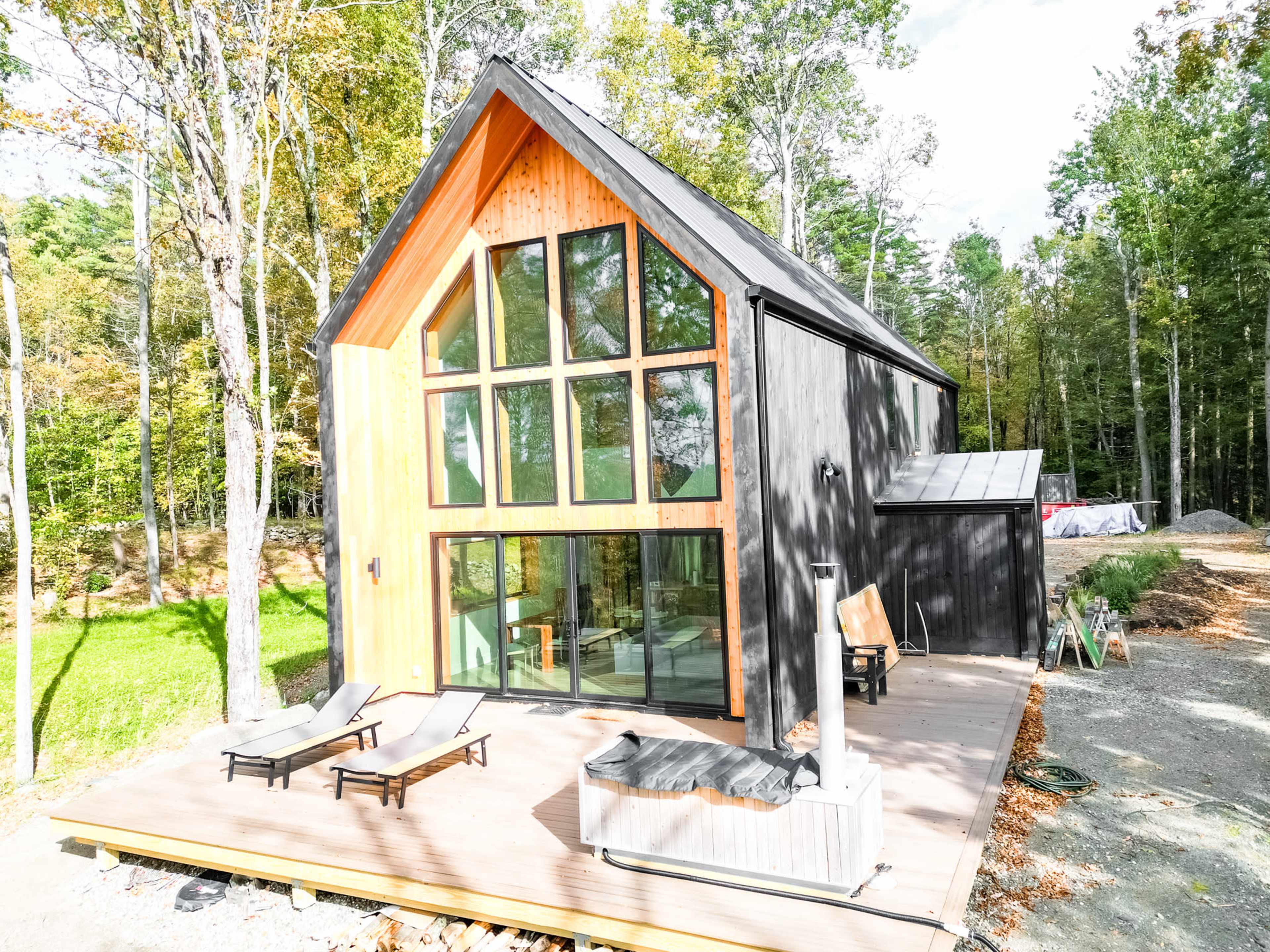 A modern, angular house with large windows and a wooden accent is situated in a wooded area, featuring a deck with lounge chairs and a hot tub.