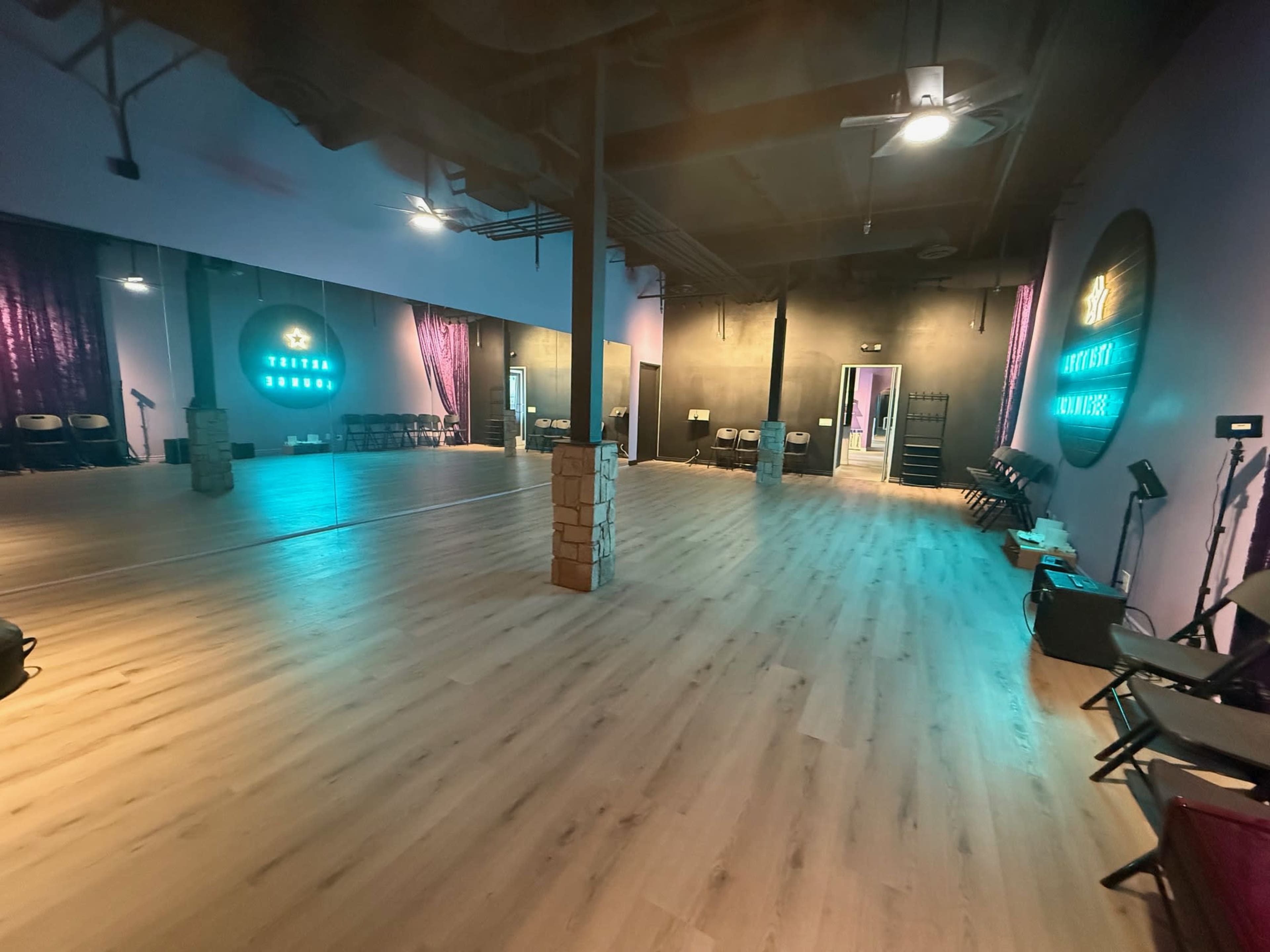 The image shows a spacious dance studio with wooden flooring, large mirrors on one wall, and purple curtains, illuminated by lights in various shades.