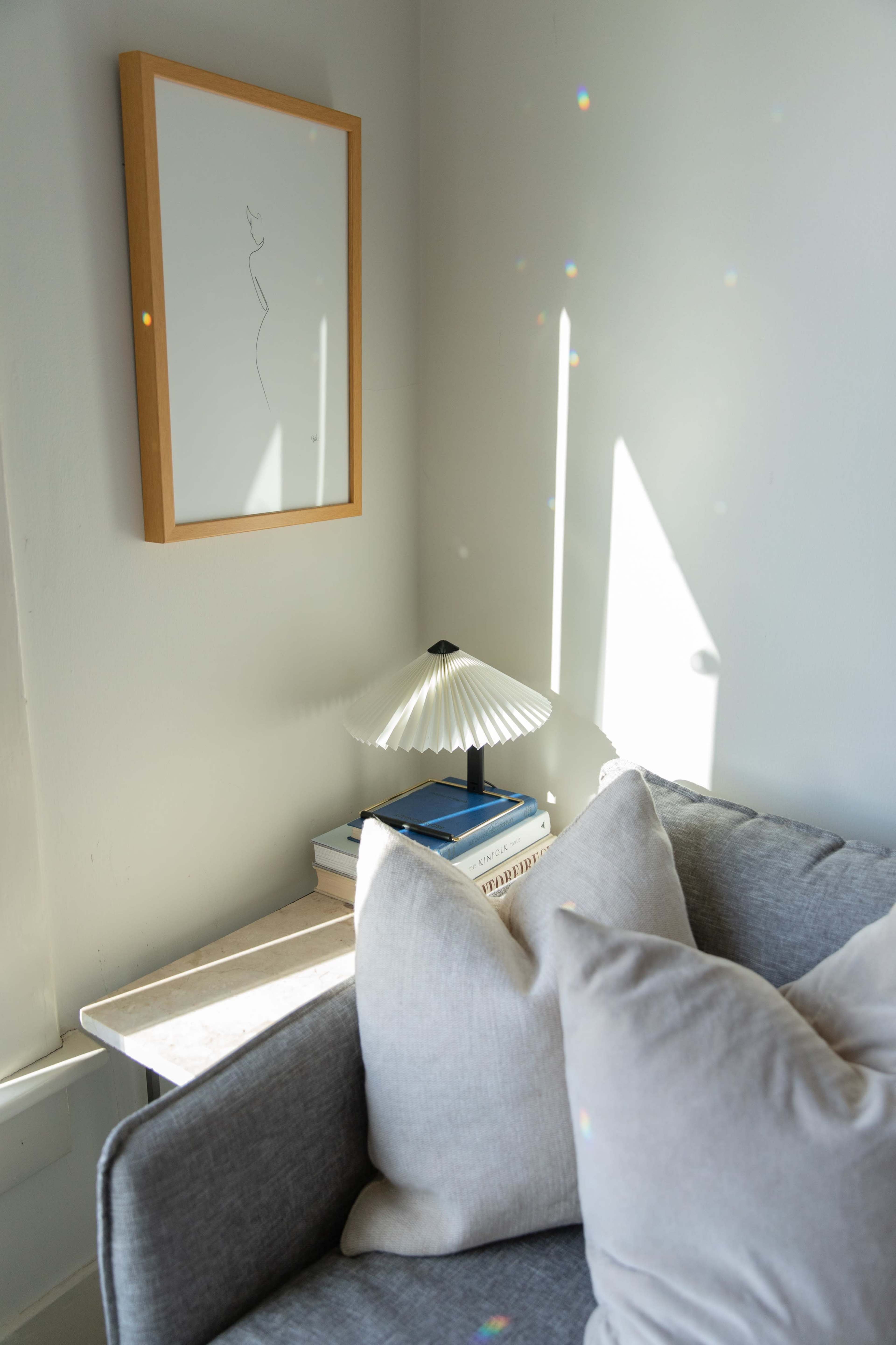 A gray sofa is positioned beside a side table with stacked books and a decorative lamp, accompanied by a framed line drawing on the wall.