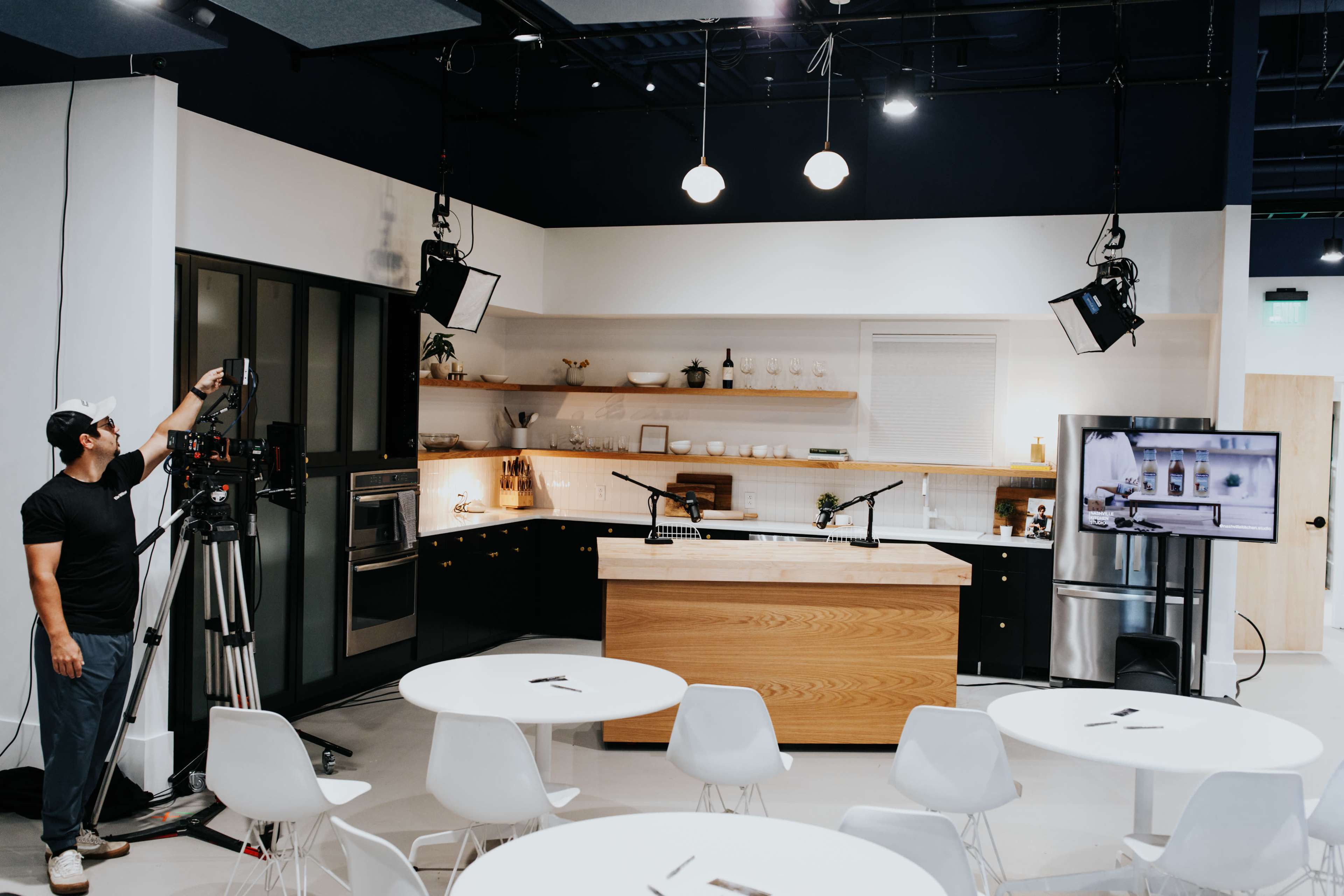 A man adjusts equipment in a modern kitchen set designed for filming, featuring a wooden island and white tables.