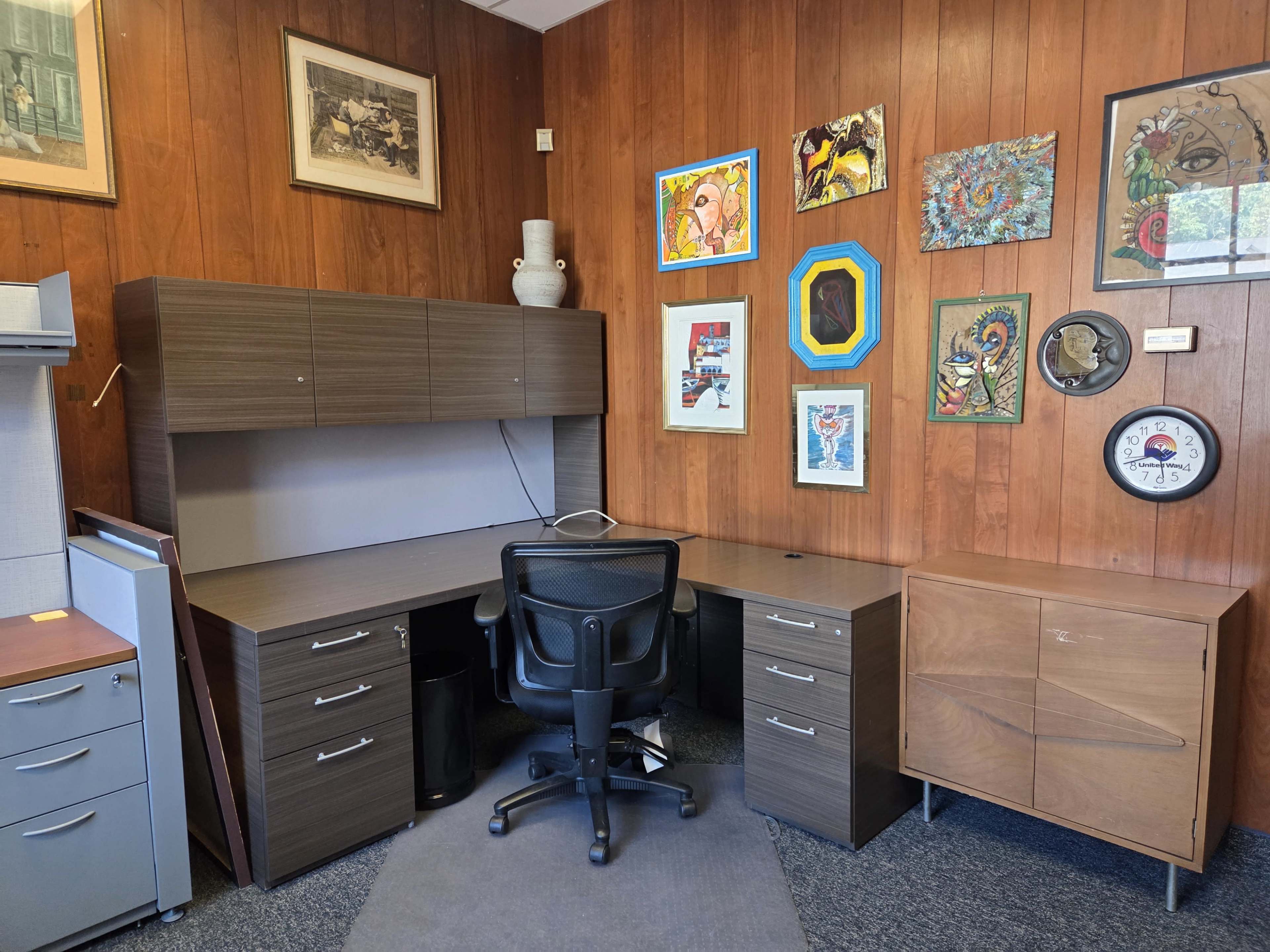 The image shows an office space featuring a desk with a chair, a filing cabinet, and a wall adorned with various framed artworks.