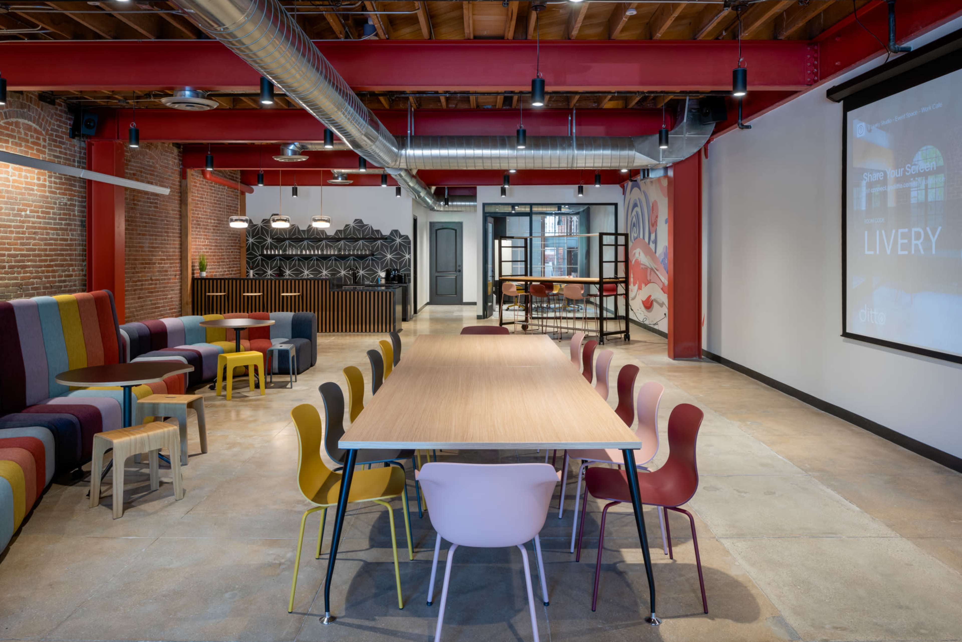 The image shows a modern open workspace with colorful seating areas, a long wooden table surrounded by chairs, and an exposed brick wall adorned with contemporary art.